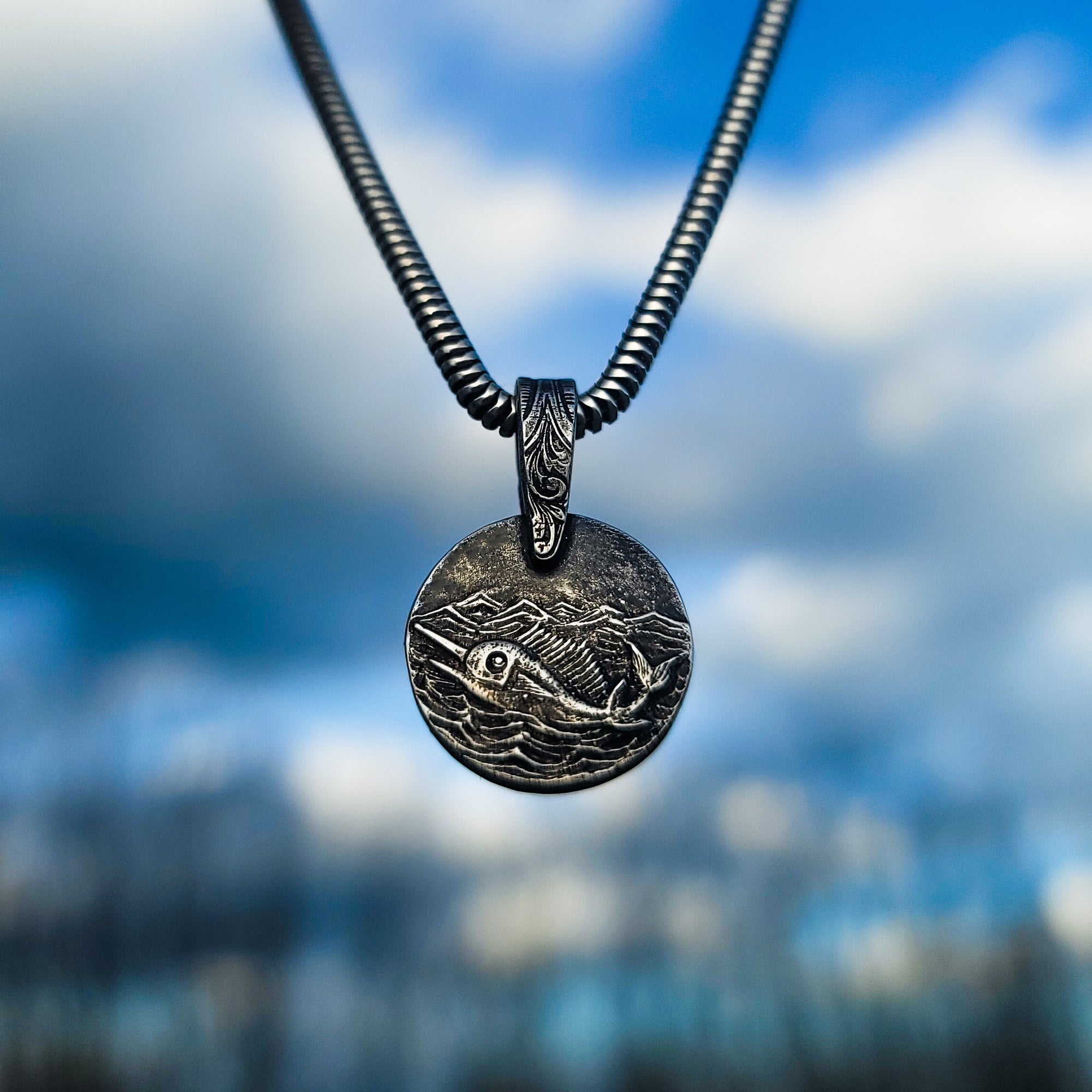 Pictured is our exquisite handmade sterling silver Swordfish in Silver Seas pendant. The pendant is suspended in front of an out-of-focus cloudy sky, creating a beautiful contrast against the intricate design of the swordfish swimming in the ocean