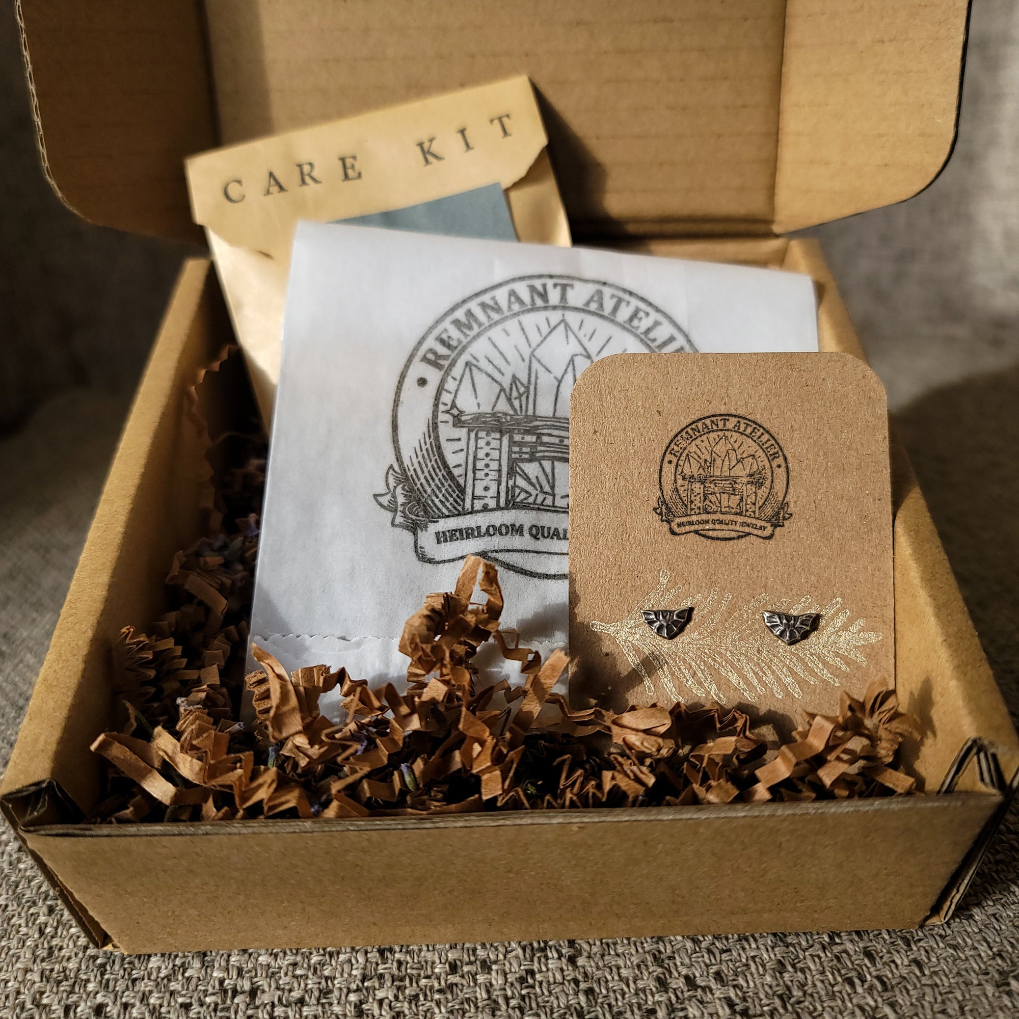 This photo shows a pair of handmade sterling silver bat-shaped stud earrings displayed on a cardboard earring card inside a cardboard box, surrounded by shredded cardboard. The package also includes a care kit to keep the jewelry in top condition.