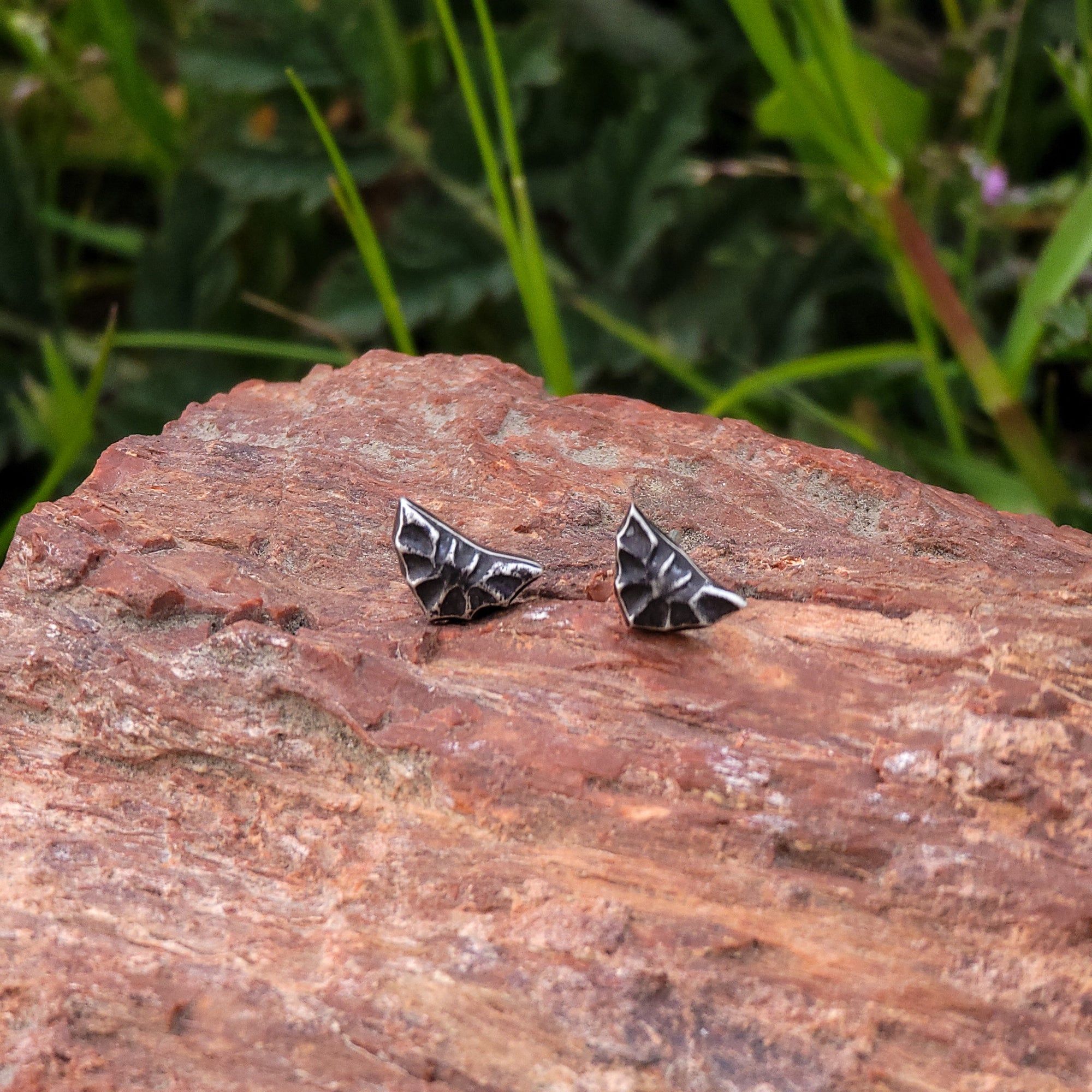 These sterling silver bat shaped stud earrings are depicted in mid-flight, with their wings extended in a graceful arc. In the photograph, the earrings are resting on a piece of wood, surrounded by lush green leaves.