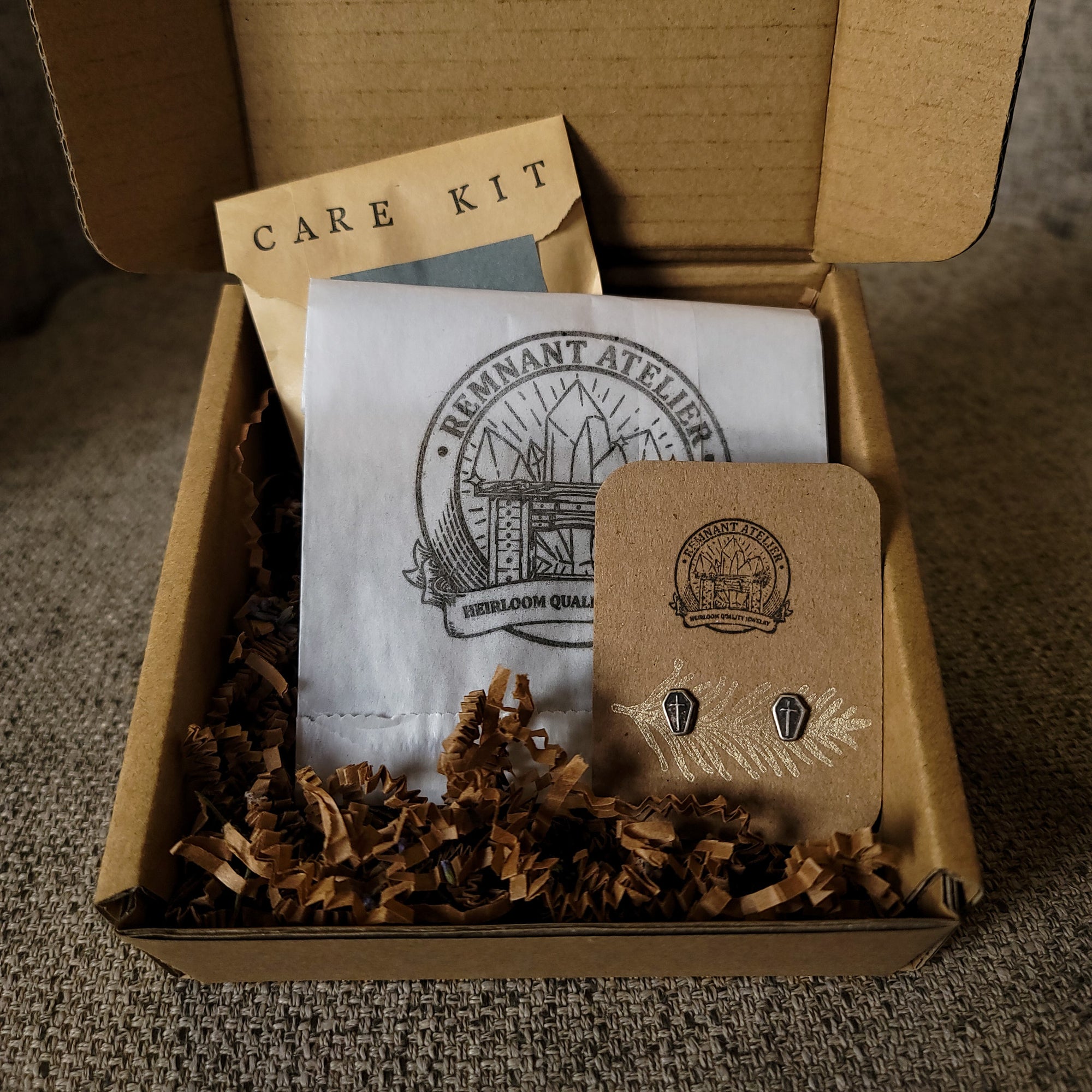 This photo shows a pair of handmade sterling silver coffin shaped stud earrings displayed on a cardboard earring card inside a cardboard box, surrounded by shredded cardboard. The package also includes a care kit to keep the jewelry in top condition.