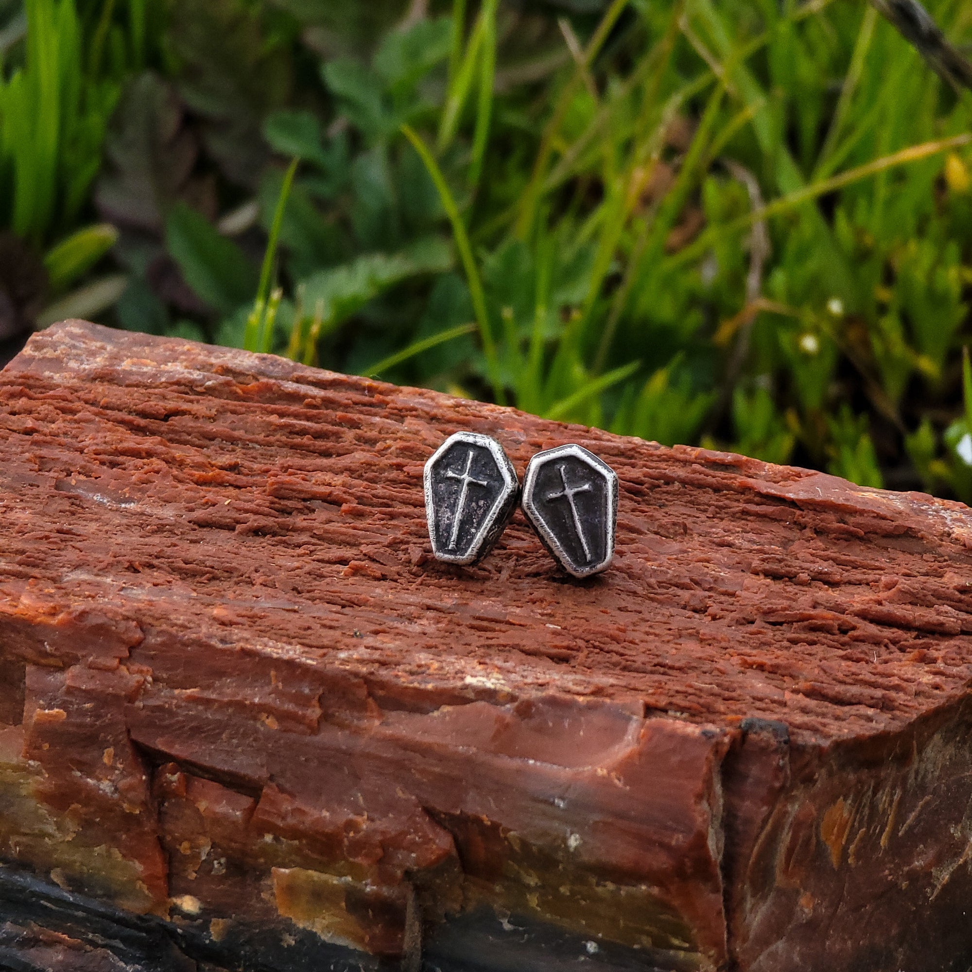 These earrings are handcrafted in the shape of small coffins. The earrings are resting on a natural piece of wood. Surrounding the earrings are lush green leaves.