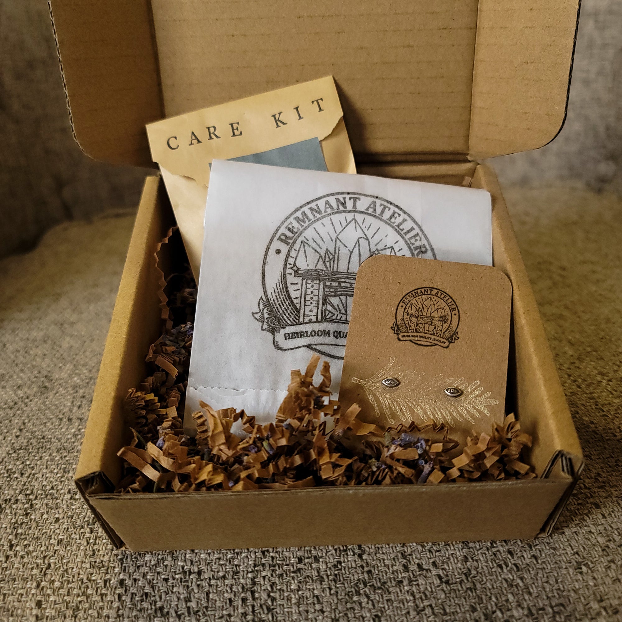 This photo shows a pair of handmade sterling silver eyes shaped stud earrings displayed on a cardboard earring card inside a cardboard box, surrounded by shredded cardboard. The package also includes a care kit to keep the jewelry in top condition.