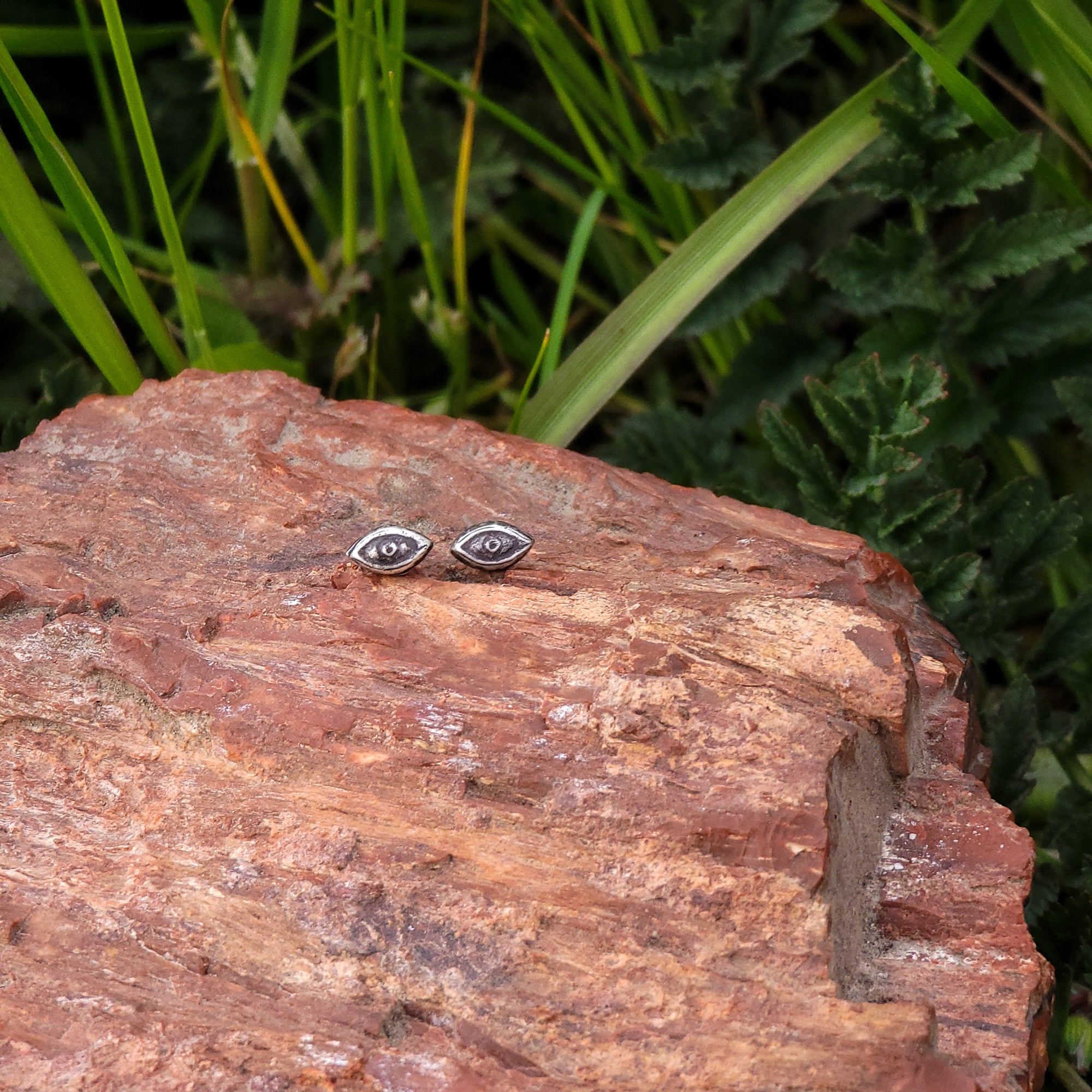 Pictured are a pair of handmade sterling silver stud earrings are designed in the shape of eyes. Placed on a wooden surface with a backdrop of green leaves, they make a statement with their unique design. Perfect for anyone who loves unique jewelry.