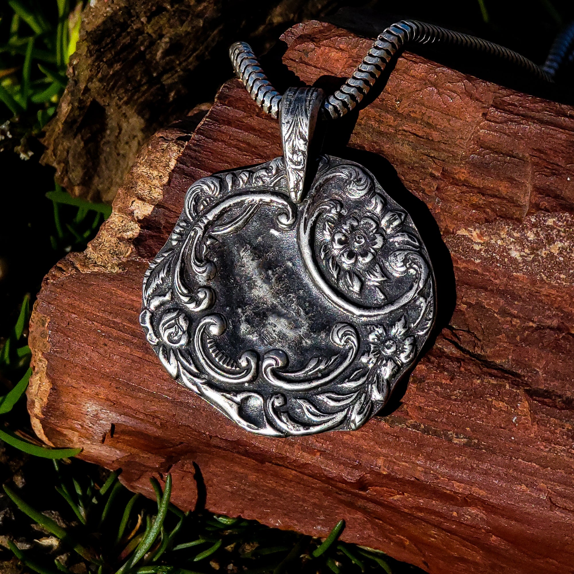 The pendant is shown resting on a piece of wood with tufts of grass visible in the background. The pendant itself is an irregular wreath of flowers and vines made from sterling silver, with a tarnished finish that brings out the details of the design