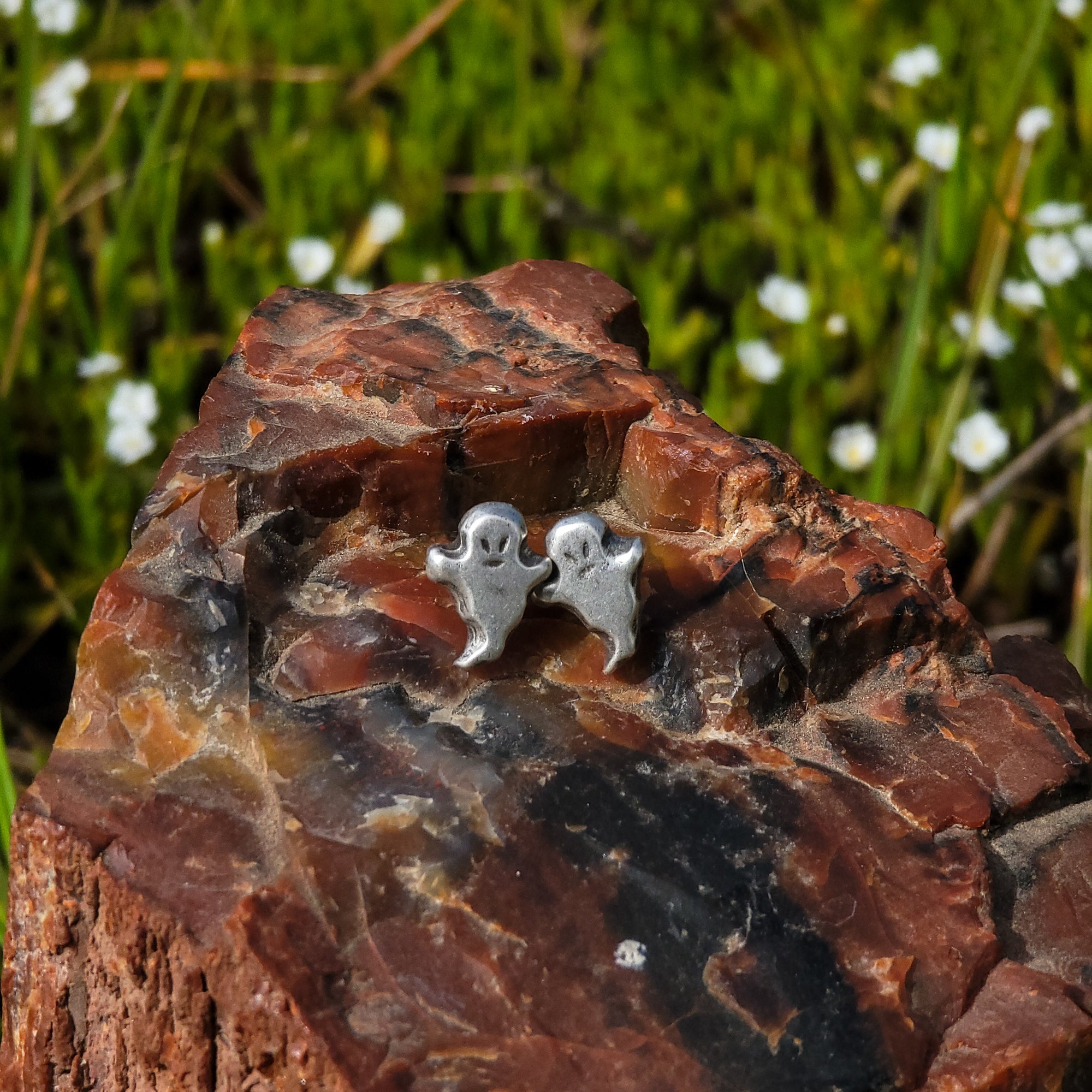 This photo features a pair of handmade sterling silver stud earrings in the shape of smiling ghosts, placed on a piece of wood with green leaves and little white flowers in the background.