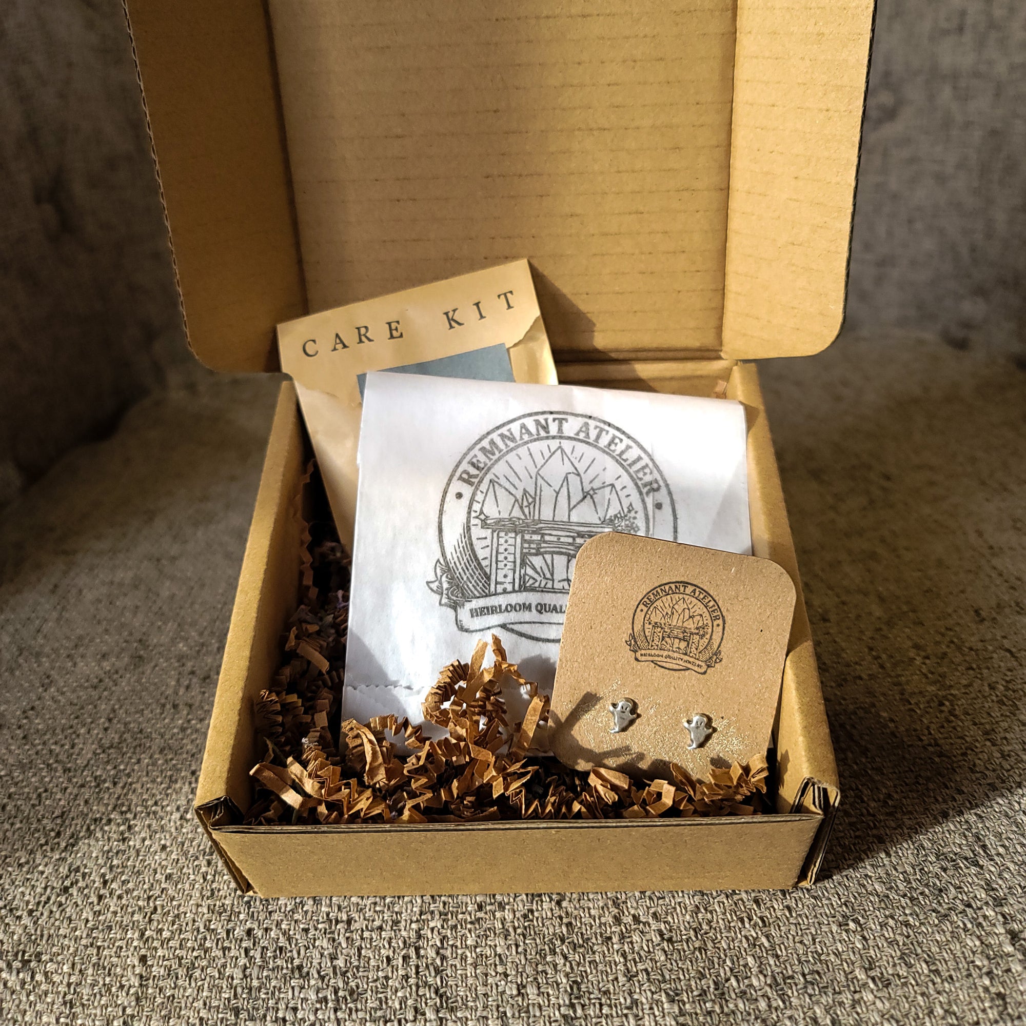 This photo shows a pair of handmade sterling silver ghost shaped stud earrings displayed on a cardboard earring card inside a cardboard box, surrounded by shredded cardboard. The package also includes a care kit to keep the jewelry in top condition.