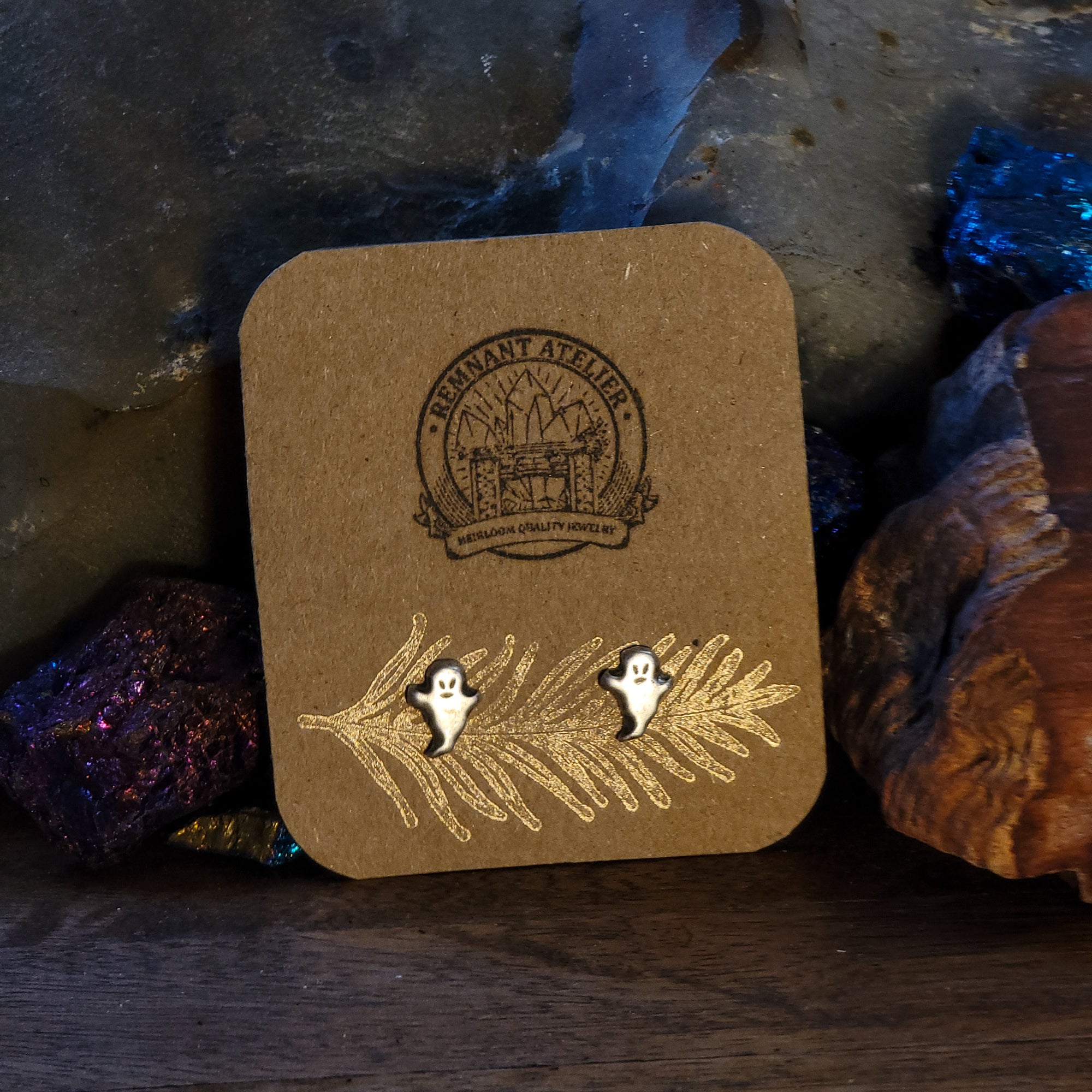 This photo features a pair of handmade sterling silver stud earrings in the shape of grinning ghosts, displayed on a cardboard earring card and surrounded by sparkling crystals.