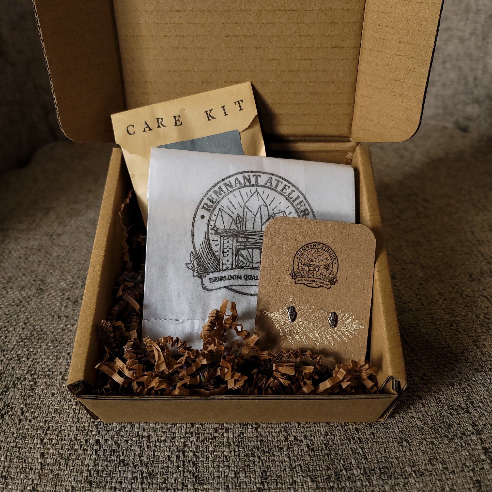 This photo shows a pair of handmade sterling silver hand shaped stud earrings displayed on a cardboard earring card inside a cardboard box, surrounded by shredded cardboard. The package also includes a care kit to keep the jewelry in top condition.