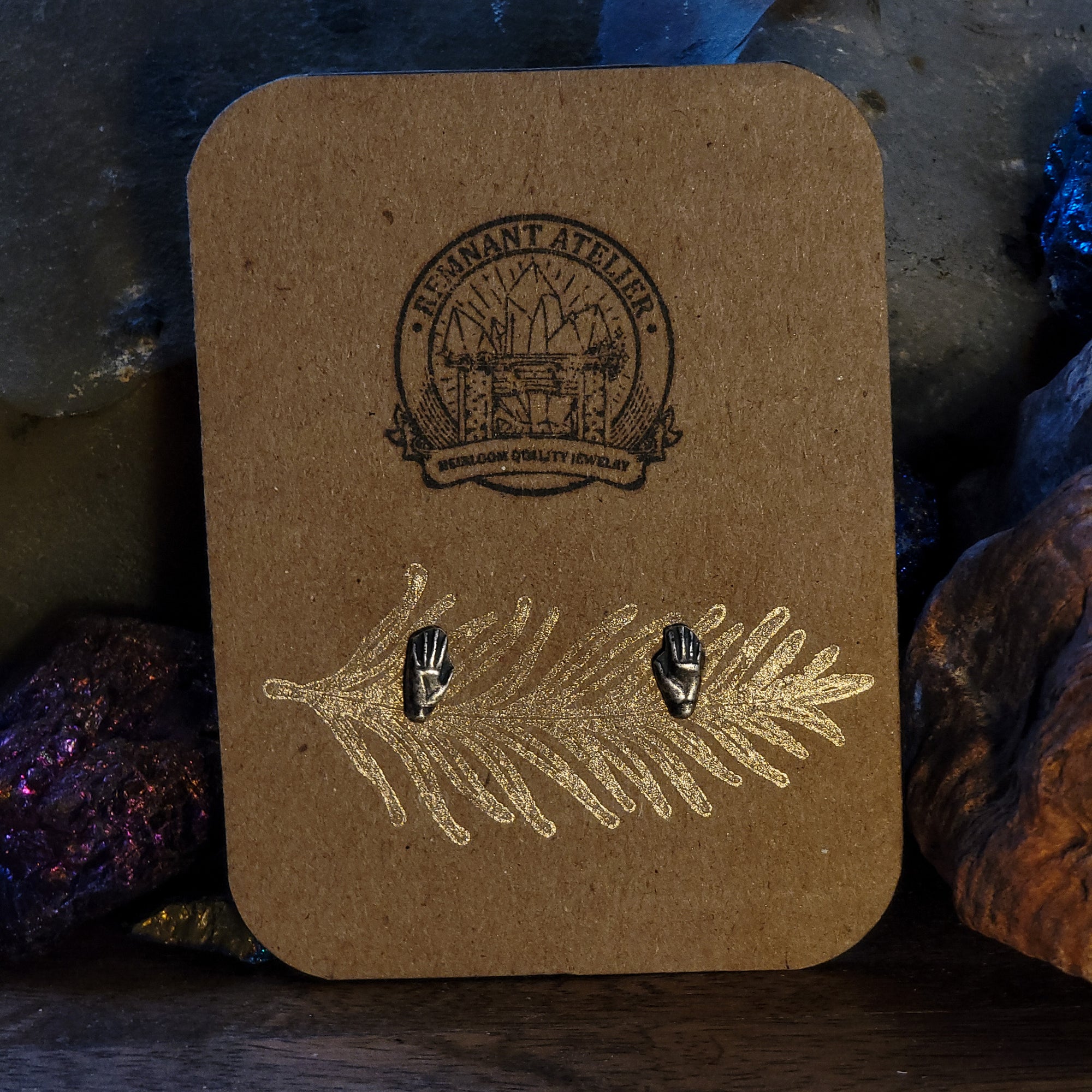 A pair of handmade sterling silver stud earrings in the shape of hands are displayed on a cardboard earring card. The intricate details of the hands are highlighted by the shine of the silver. The earrings are surrounded by sparkling crystals.