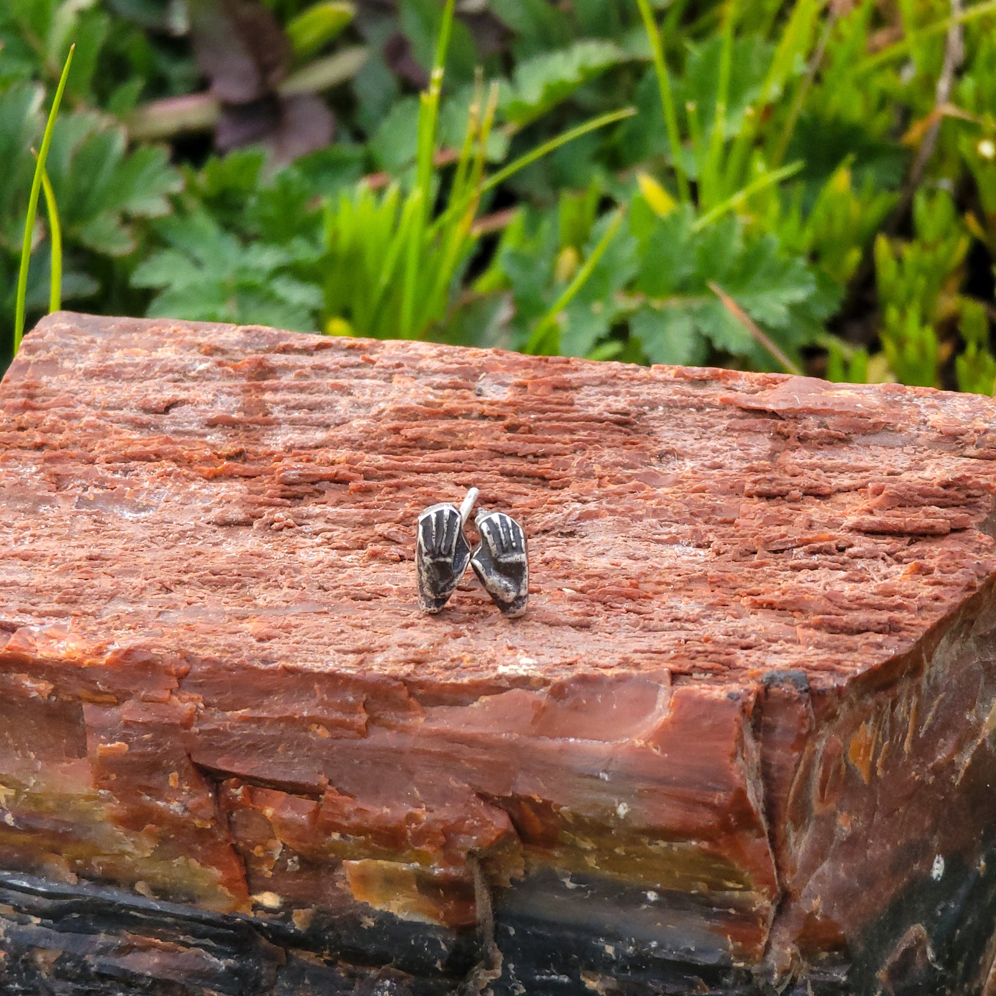 A pair of handmade sterling silver stud earrings in the shape of hands are displayed on a piece of wood. The details of the hands, including the fingers and palms, are delicately crafted. In the background, there are lush green leaves.
