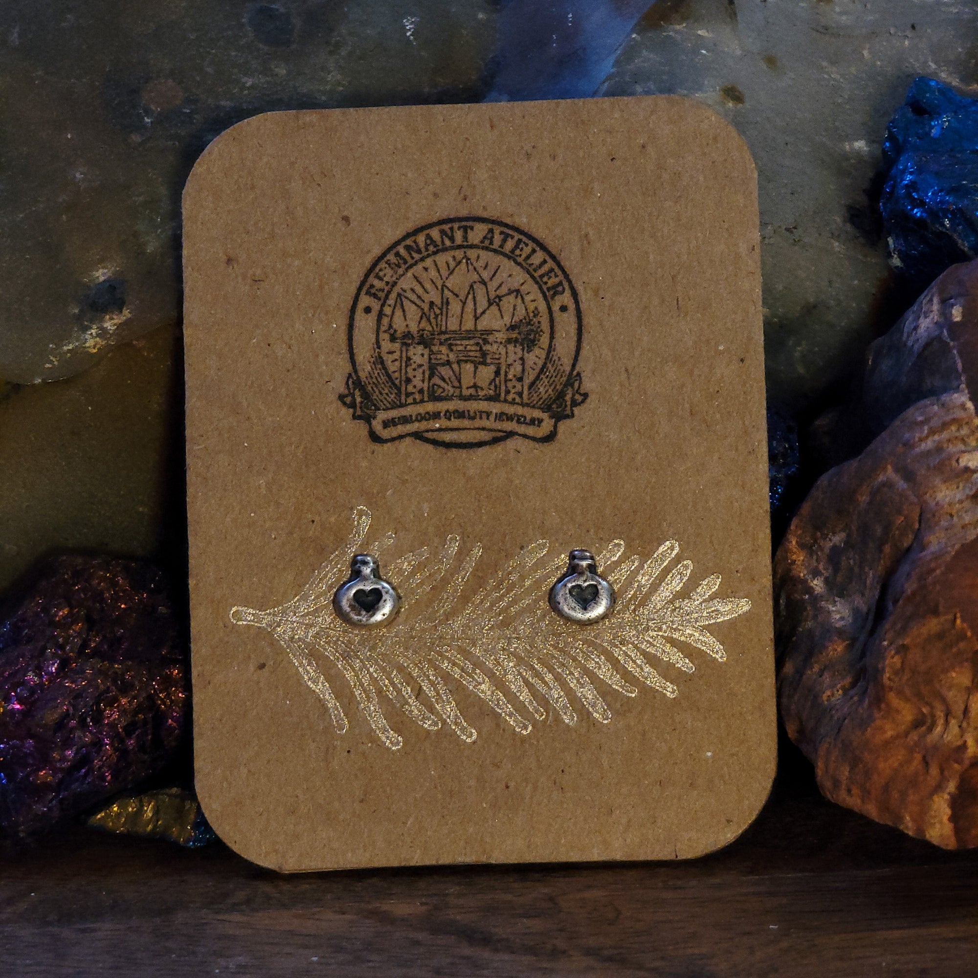 A pair of handmade sterling silver stud earrings in the shape of love potions are displayed on a cardboard earring card, resting upon glittering crystals. A unique and enchanting piece of jewelry perfect for those who believe in the power of love.