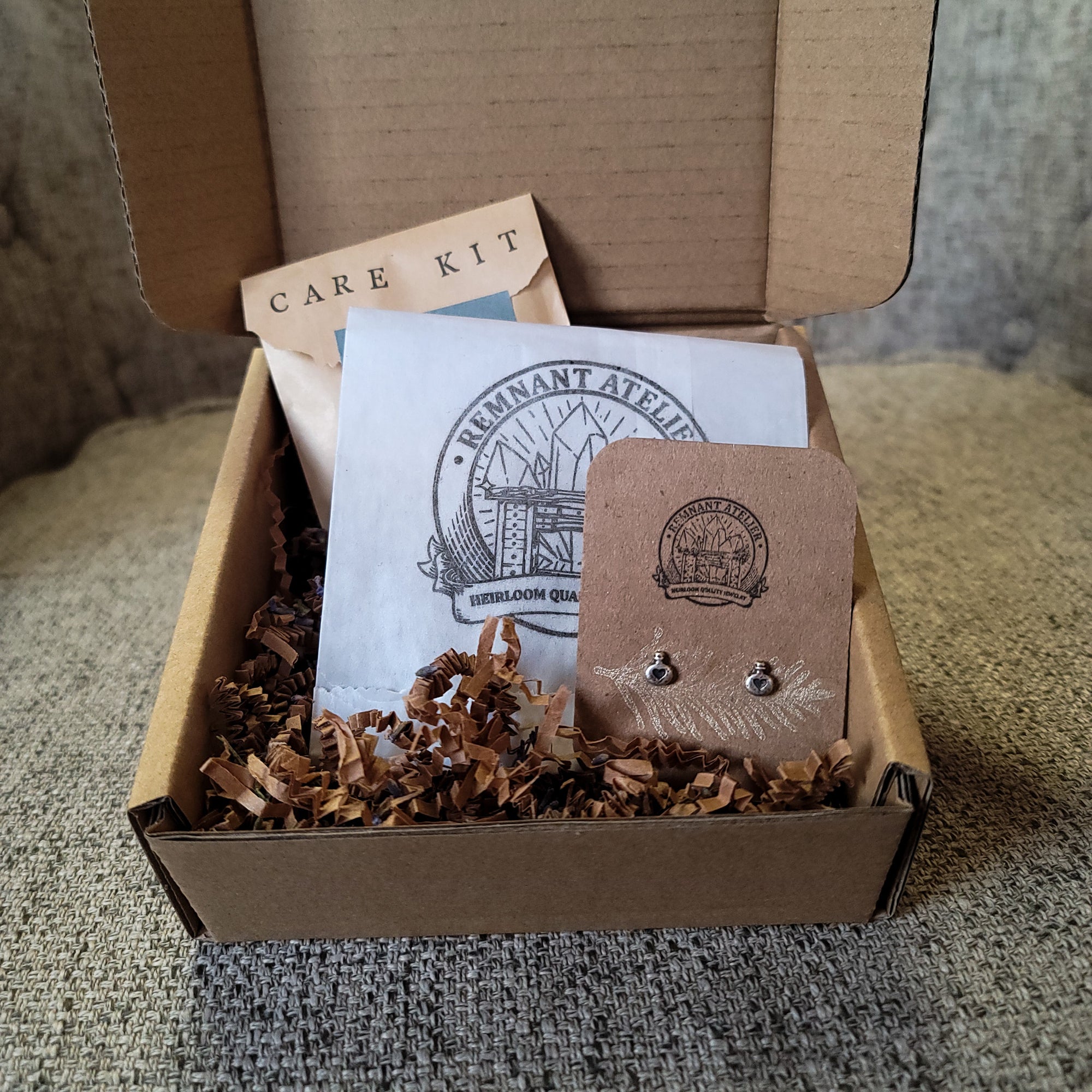 This photo shows a pair of handmade sterling silver love potion shaped stud earrings displayed on a cardboard earring card inside a box, surrounded by shredded cardboard. The package also includes a care kit to keep the jewelry in top condition.