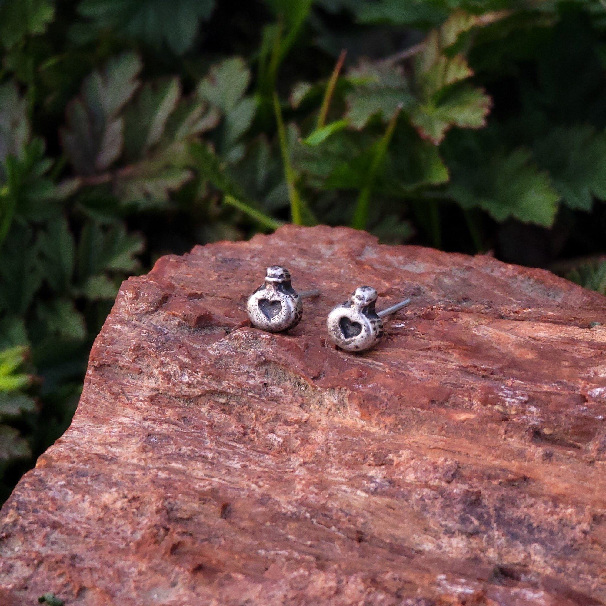 A pair of handmade sterling silver stud earrings in the shape of love potions are displayed on a piece of wood. In the background, there are green leaves. The earrings are intricately crafted to resemble potion bottles with a heart shape label.