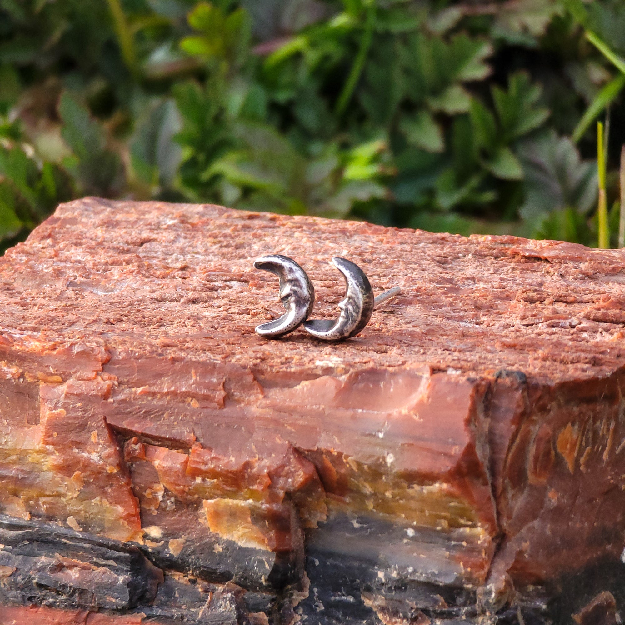 A pair of handmade sterling silver stud earrings in the shape of smiling moons are displayed on a piece of wood. The moons have a crescent shape and a gentle smile. In the background, there are lush green leaves that add a touch of nature.