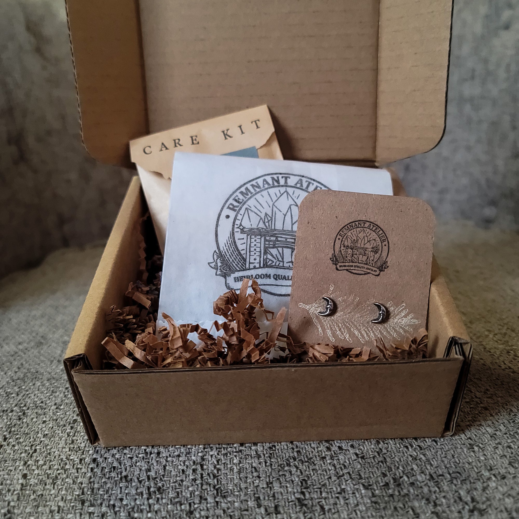 This photo shows a pair of handmade sterling silver moon shaped stud earrings displayed on a cardboard earring card inside a cardboard box, surrounded by shredded cardboard. The package also includes a care kit to keep the jewelry in top condition.