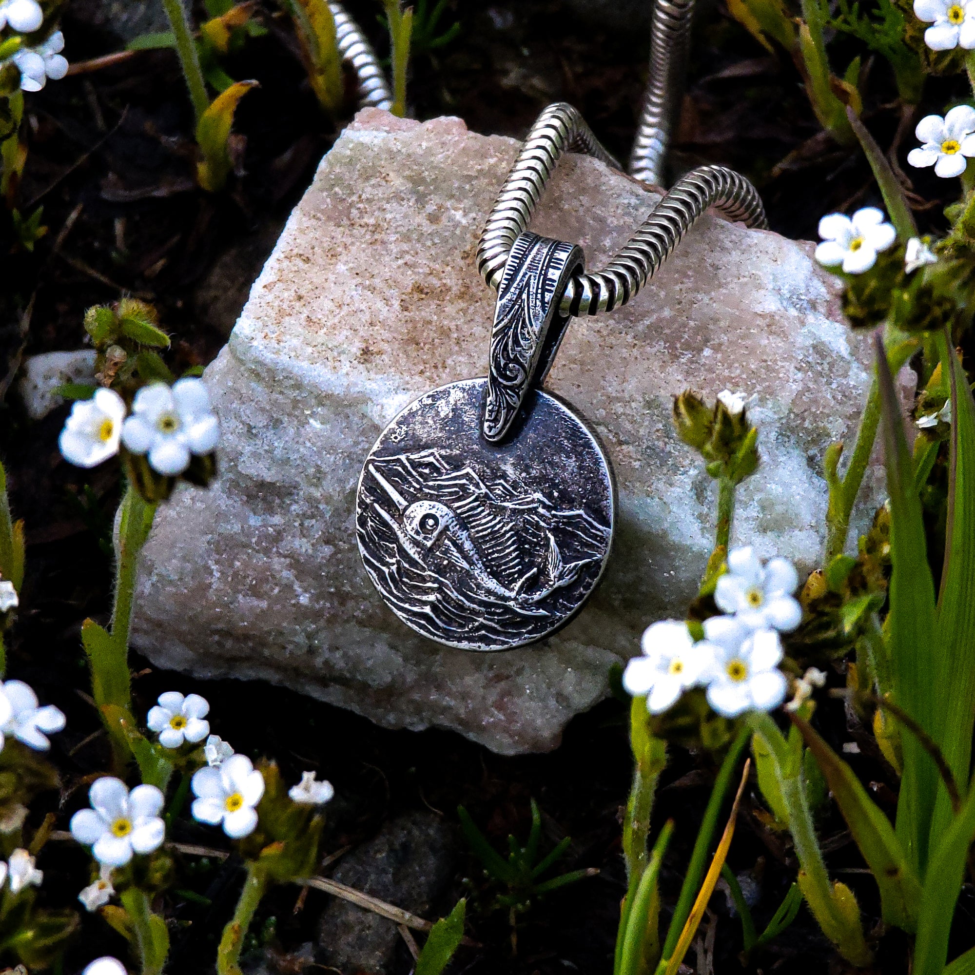 This photo showcases our handmade solid sterling silver Swordfish in Silver Seas pendant. The detailed design features a swordfish swimming in a stormy ocean. The pendant is resting on a beige rock and surrounded by tiny white flowers.