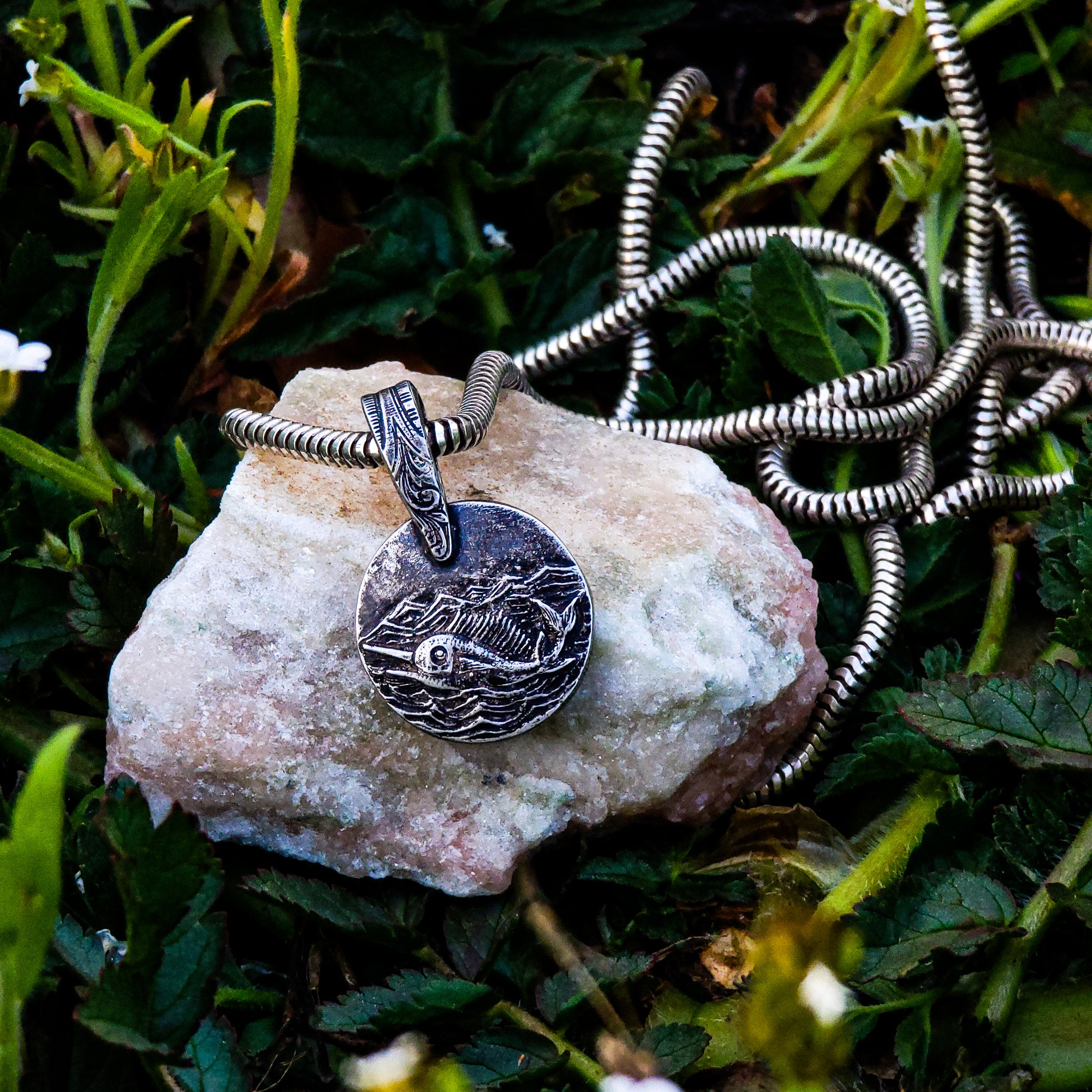 This photo showcases our stunning handmade solid sterling silver Swordfish in Silver Seas pendant. The pendant features a swordfish swimming in the ocean under a stormy sky. The pendant is resting on a beige rock and surrounded by lush green foliage.