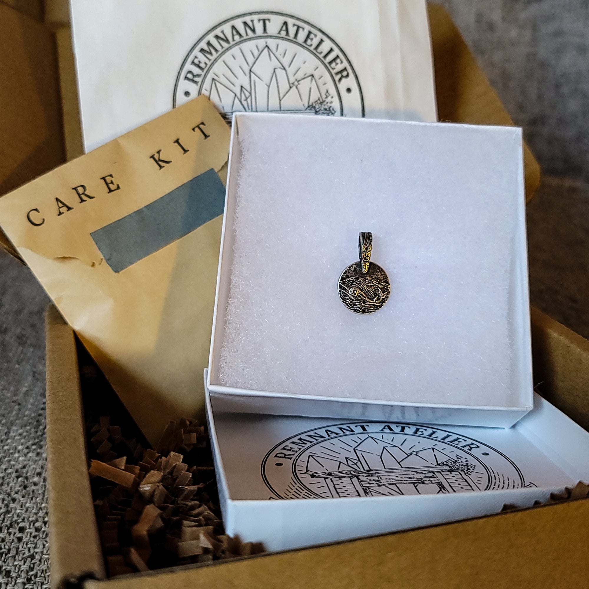 This photo shows a handmade sterling silver pendant featuring a swordfish swimming in the ocean. The pendant is placed inside a white gift box placed inside a cardboard box. The package also includes a care kit to keep the jewelry in top condition.