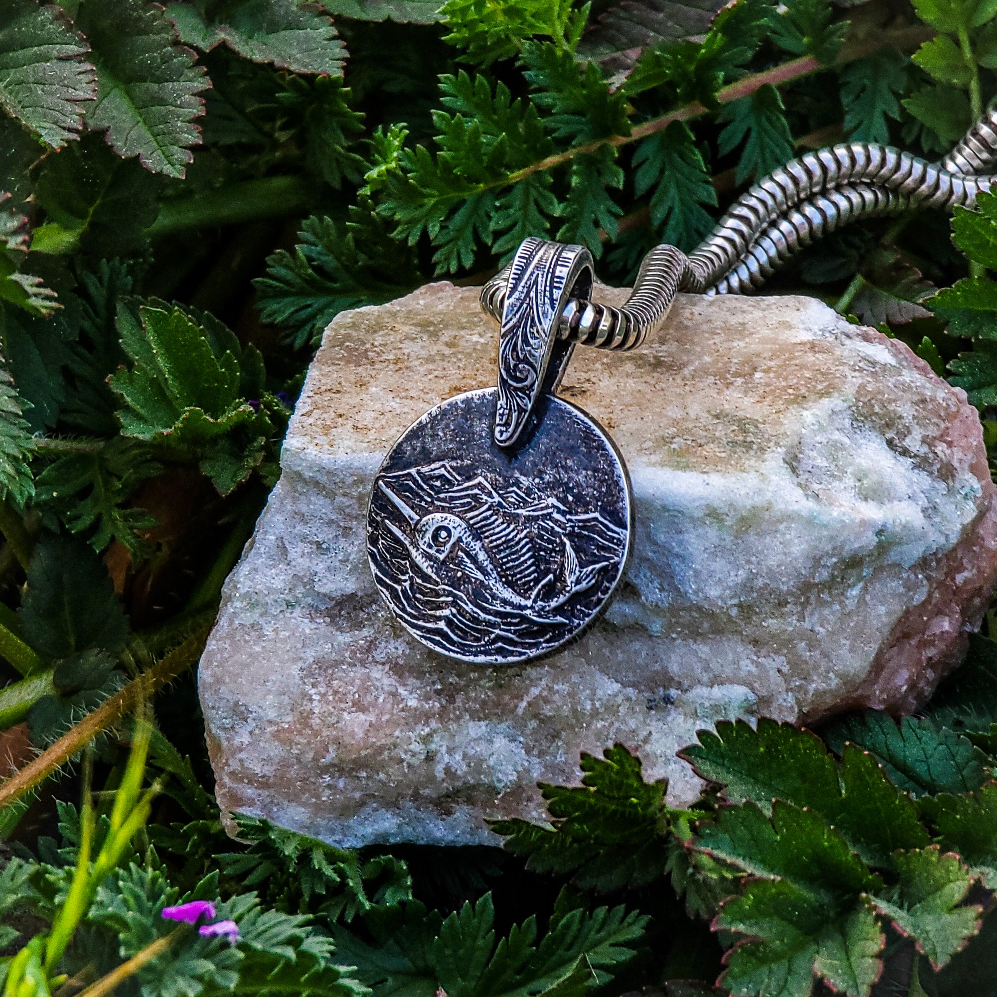 This photo showcases our stunning handmade solid sterling silver Swordfish in Silver Seas pendant. The pendant features a swordfish swimming in the ocean under a stormy sky. The pendant is resting on a beige rock and surrounded by lush green foliage.