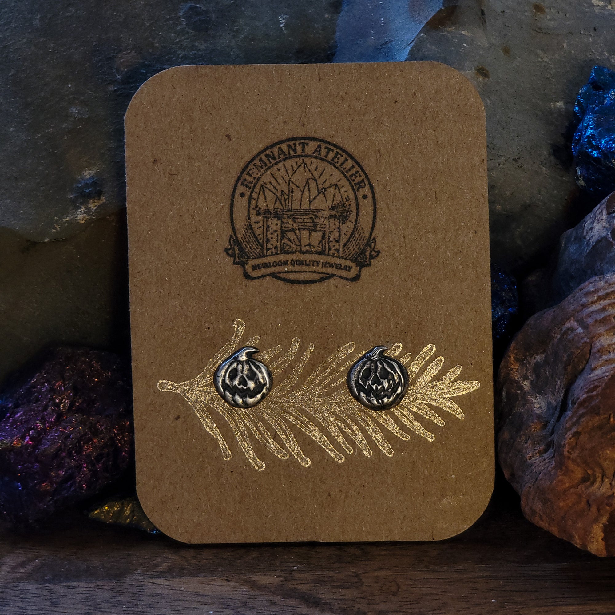 A pair of handmade sterling silver stud earrings shaped like jack o' lantern pumpkins sit on a cardboard earring card. The earrings are surrounded by sparkling crystals. The lighting shows the intricate details of the grinning pumpkins faces.