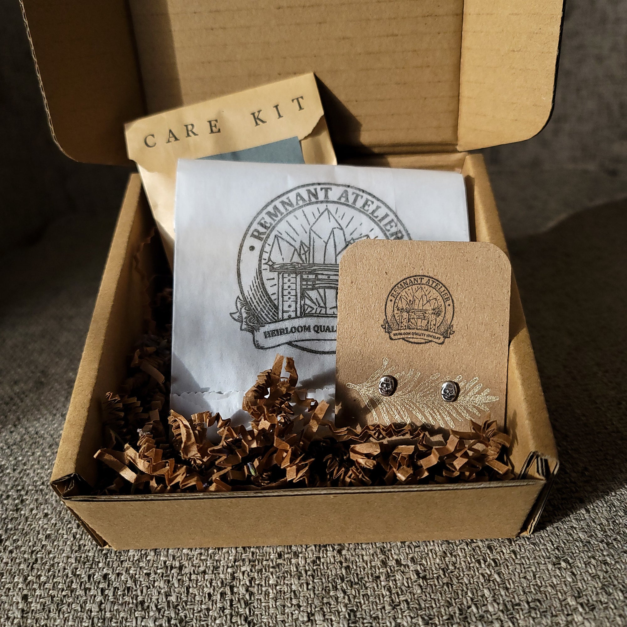 This photo shows a pair of handmade sterling silver skull shaped stud earrings displayed on a cardboard earring card inside a cardboard box, surrounded by shredded cardboard. The package also includes a care kit to keep the jewelry in top condition.