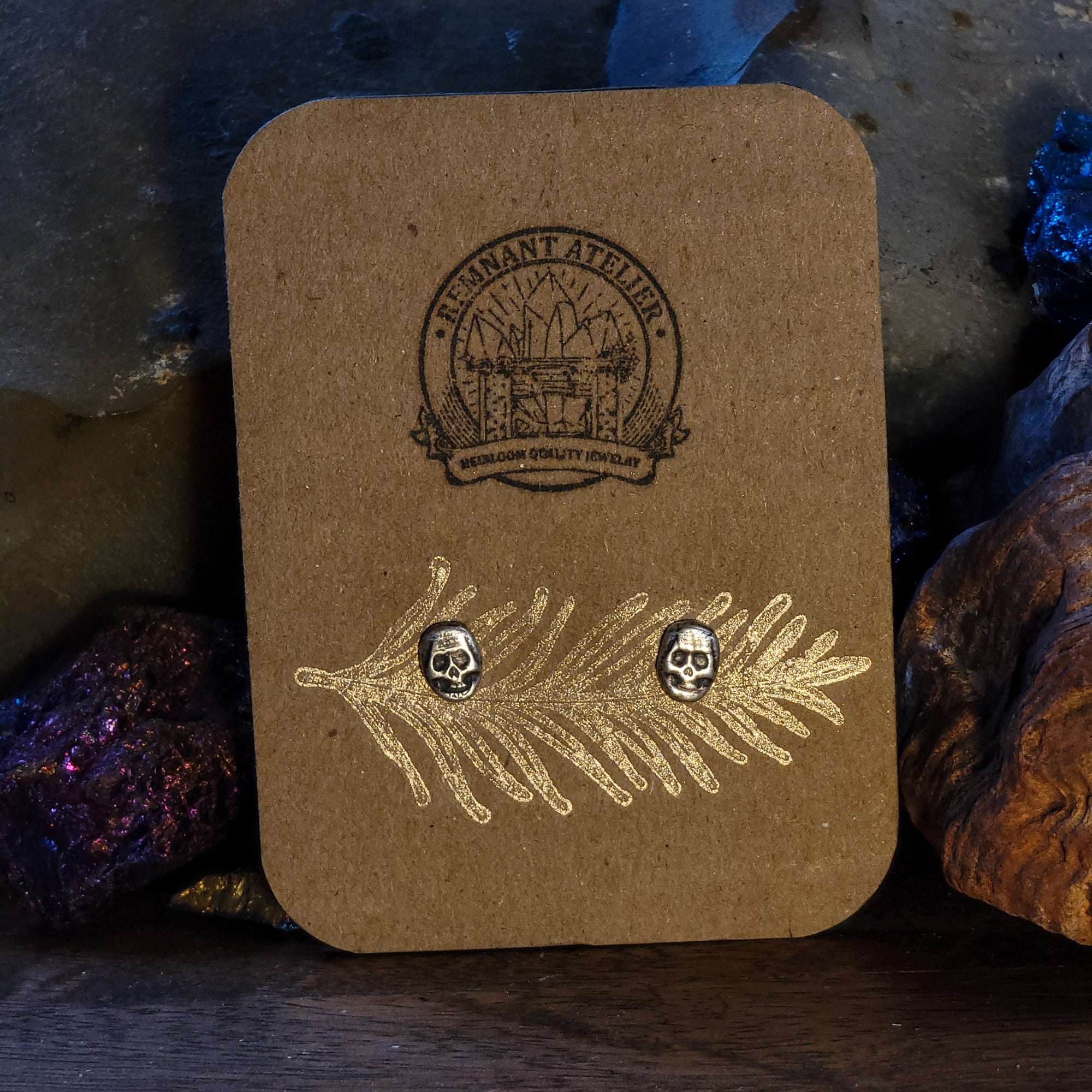 A pair of handmade sterling silver stud earrings in the shape of skulls are displayed on a cardboard earring card, resting on glittering crystals. A unique and striking piece of jewelry perfect for those who love the gothic aesthetic.