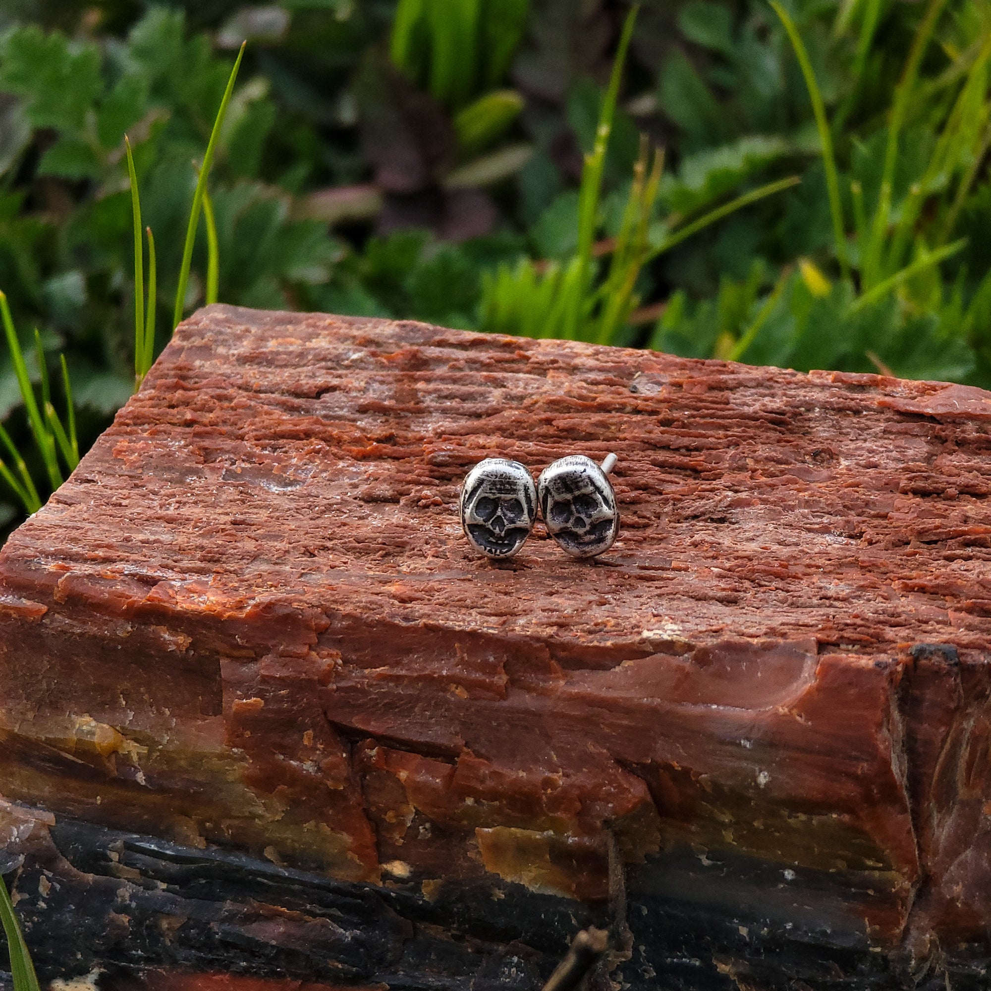 A pair of handmade sterling silver stud earrings in the shape of skulls are displayed on a piece of wood. In the background, there are lush green leaves. The intricate details of the skull design are bold and edgy.