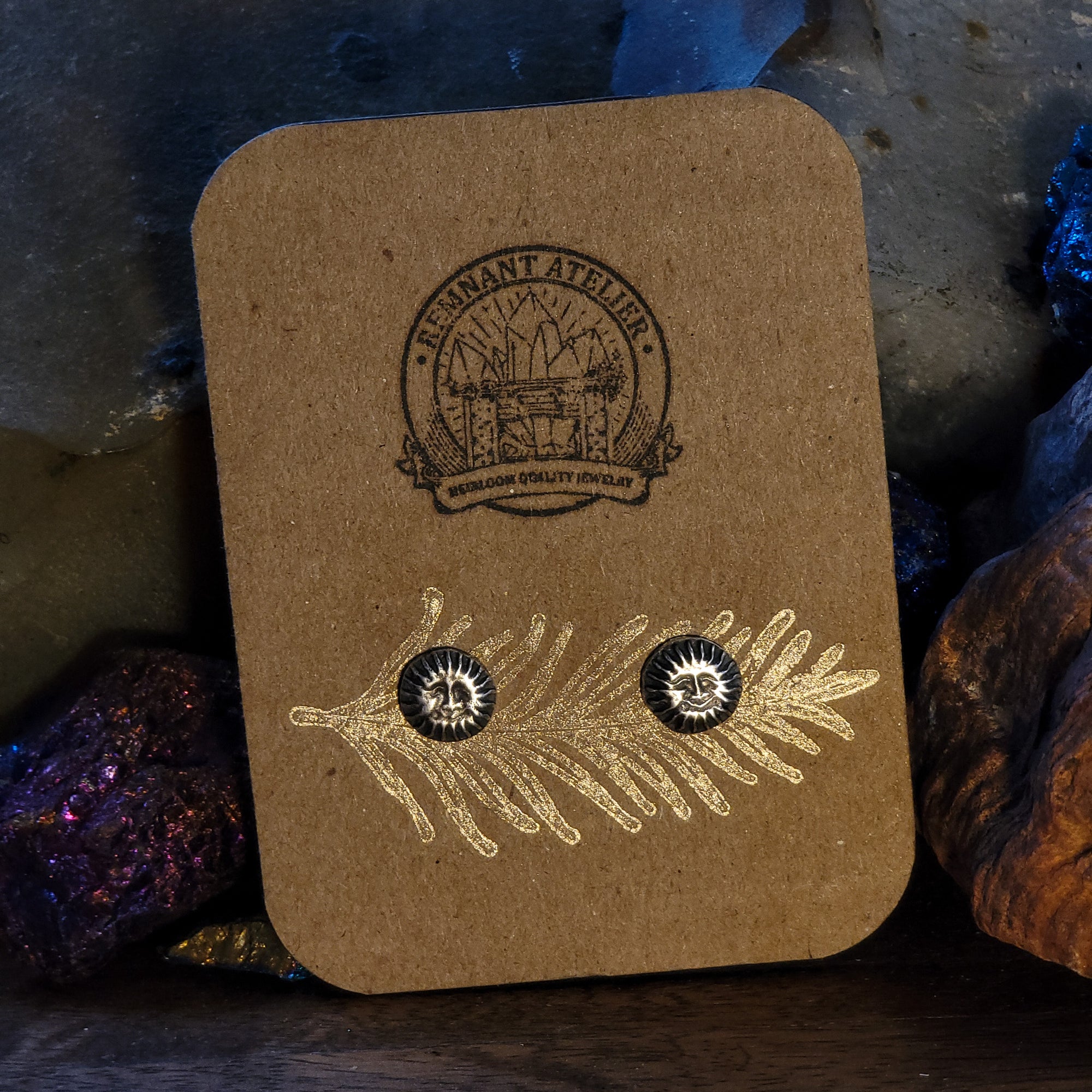 A pair of handmade sterling silver stud earrings in the shape of smiling suns are displayed on a cardboard earring card, surrounded by glittering crystals. The intricate details of the sun create a bright and cheerful look.