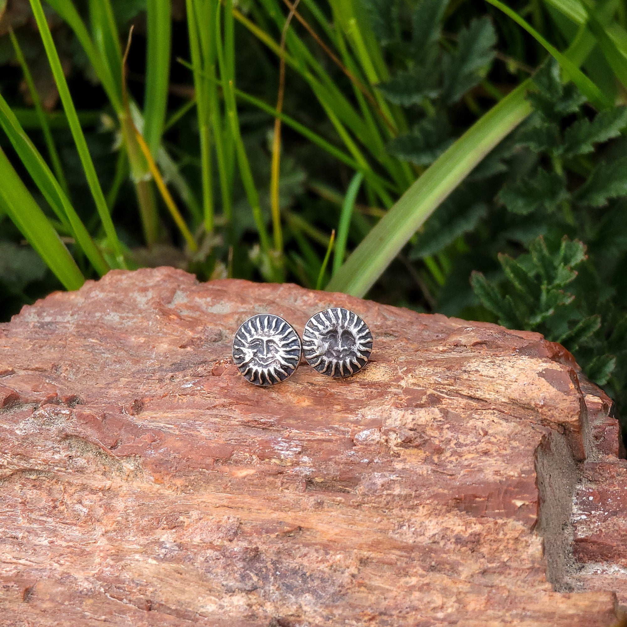 A pair of handmade sterling silver stud earrings in the shape of grinning suns are displayed on a piece of wood, with green leaves in the background. A unique and beautiful piece of jewelry perfect for those who love the warmth and energy of the sun.