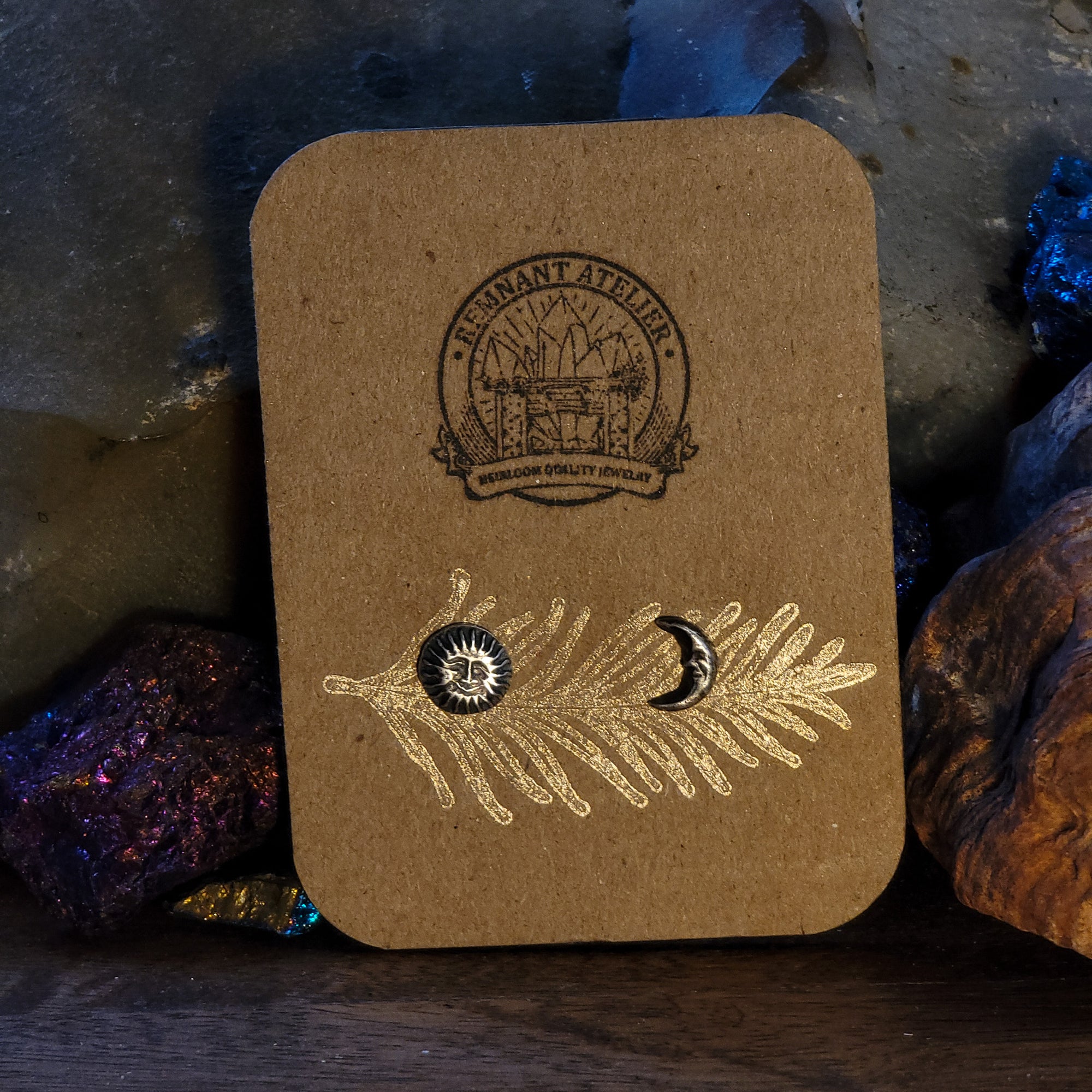 A pair of handmade sterling silver stud earrings shaped like a sun and moon are displayed on a cardboard earring card. The earring card is leaning on crystals. The intricate details of the celestial symbols are highlighted by the shine of the silver