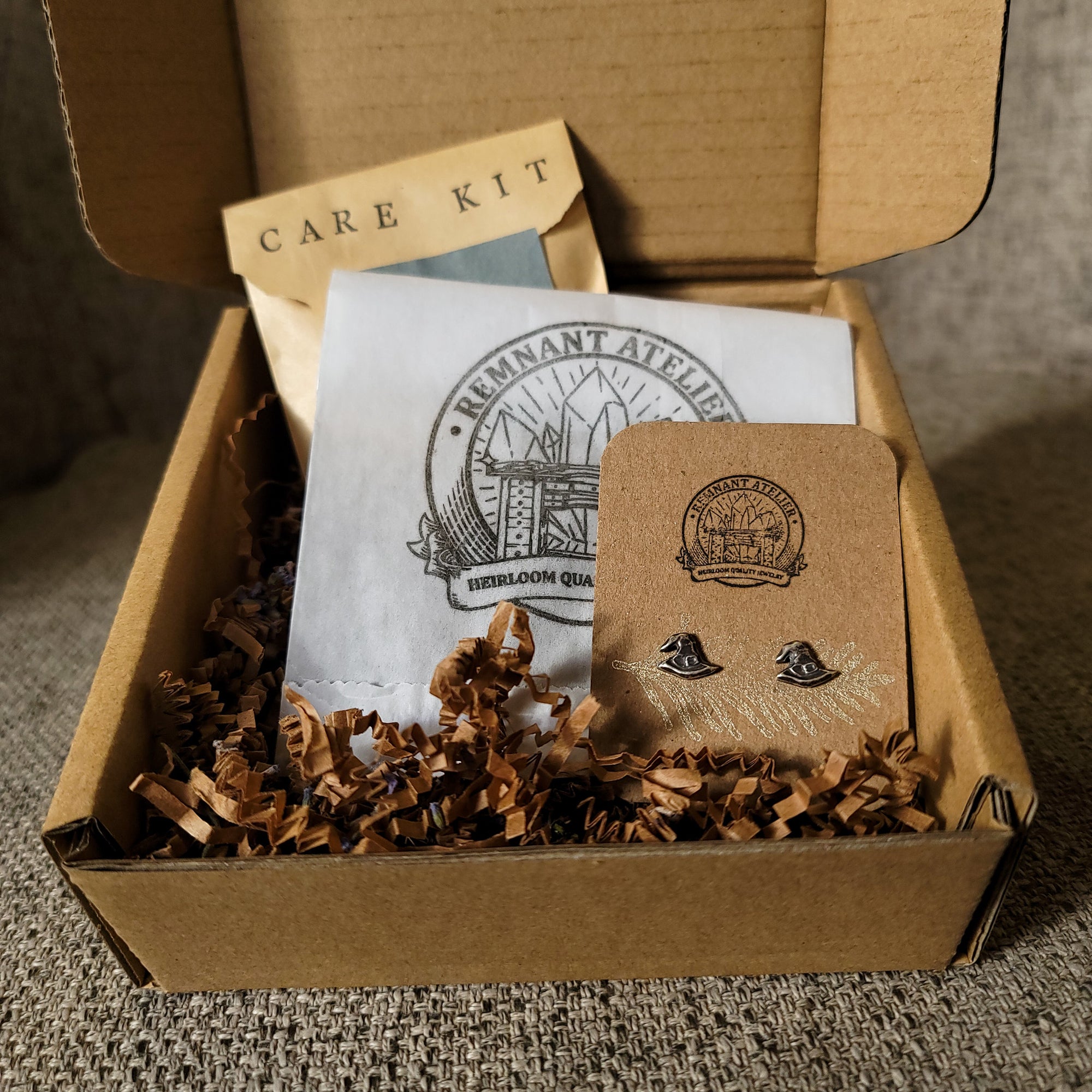 This photo shows a pair of handmade sterling silver witch hat shaped stud earrings displayed on a cardboard earring card inside a box, surrounded by shredded cardboard. The package also includes a care kit to keep the jewelry in top condition
