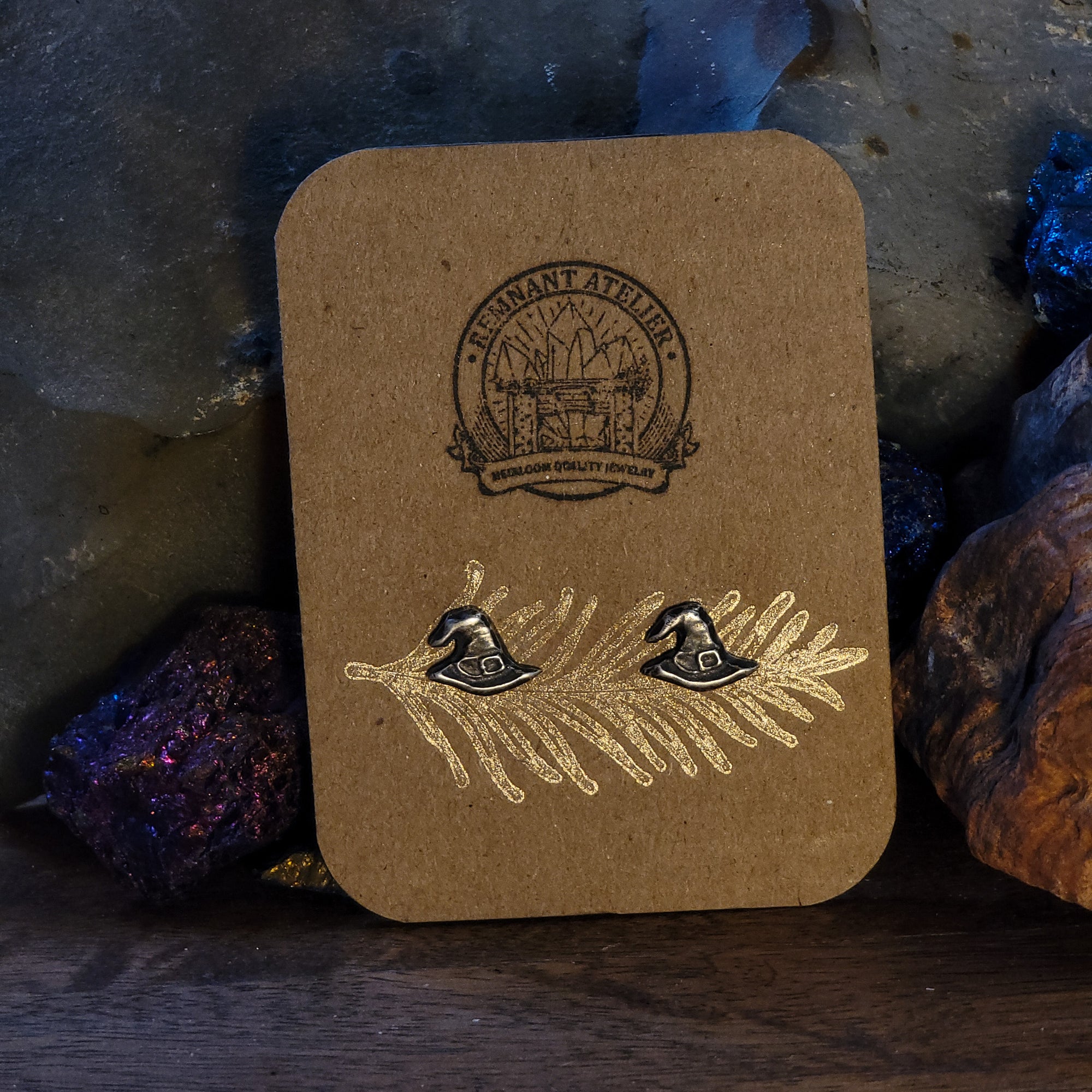 A pair of handmade sterling silver stud earrings in the shape of witch hats are displayed on a cardboard earring card, surrounded by crystals. A perfect piece of jewelry for those who love Halloween or witchy aesthetics.