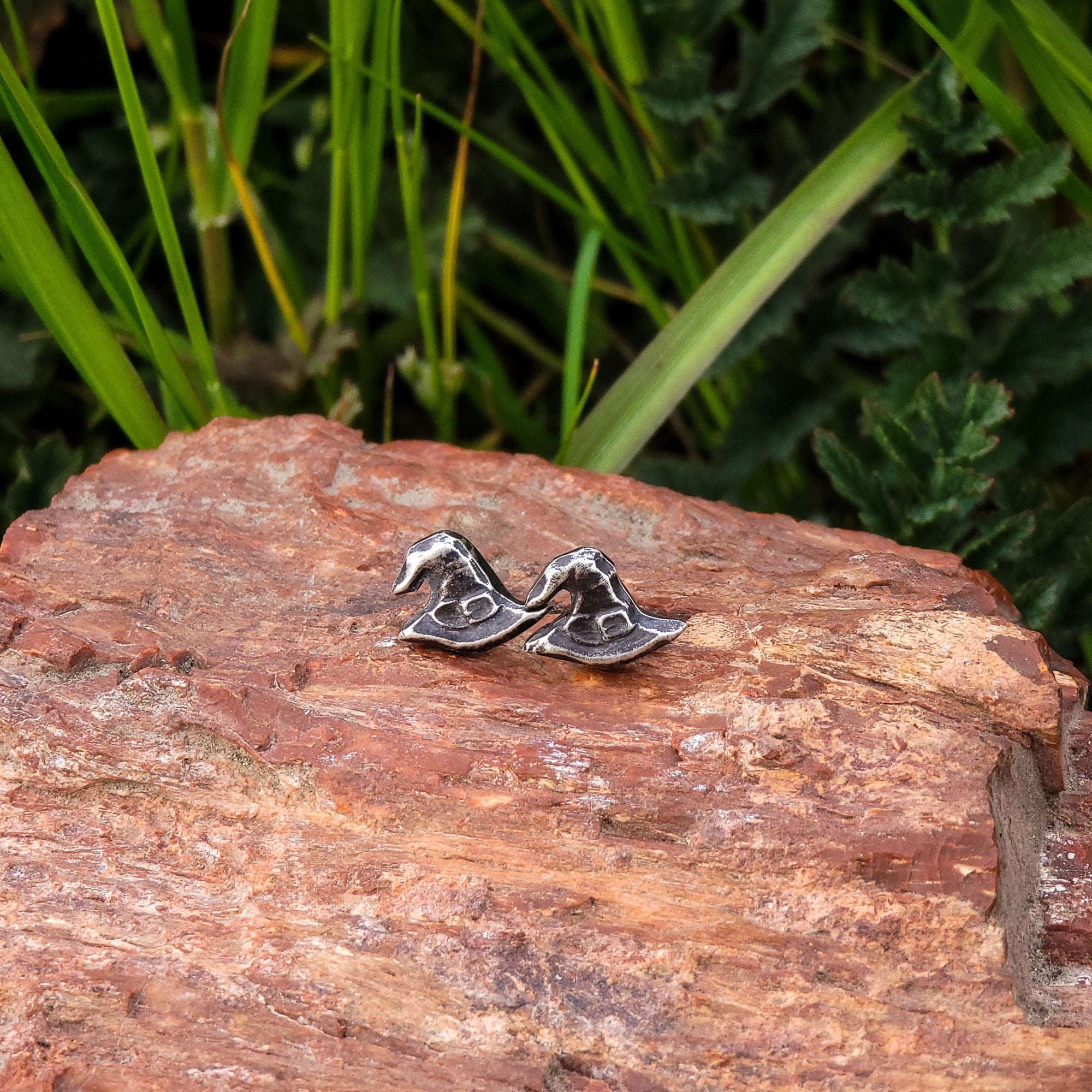 A pair of handmade sterling silver stud earrings in the shape of witch hats are displayed on a piece of wood, with green leaves in the background. The intricate details of the hat design are skillfully crafted, creating a whimsical and spooky look.