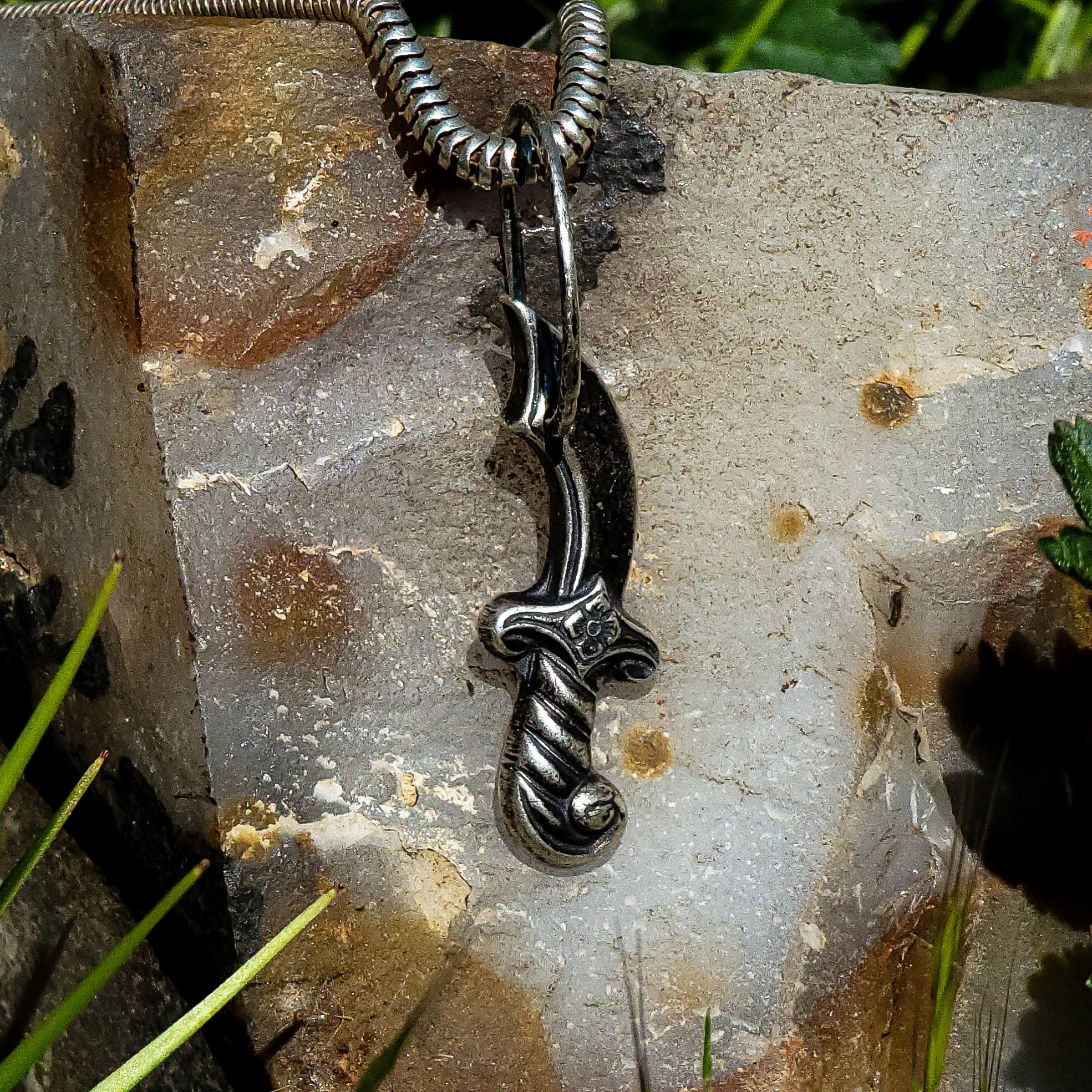 In the center of the image is a handmade sterling silver pendant in the shape of an ornate dagger with a unique tarnished finish. The pendant is resting on a large, flat rock with a multicolored beige pattern. In the background of the image, tufts of grass can be seen. The pendant's hammer detailed jump ring and the tarnished finish are accentuated by the neutral background of the rock, which appears to have a rough texture.