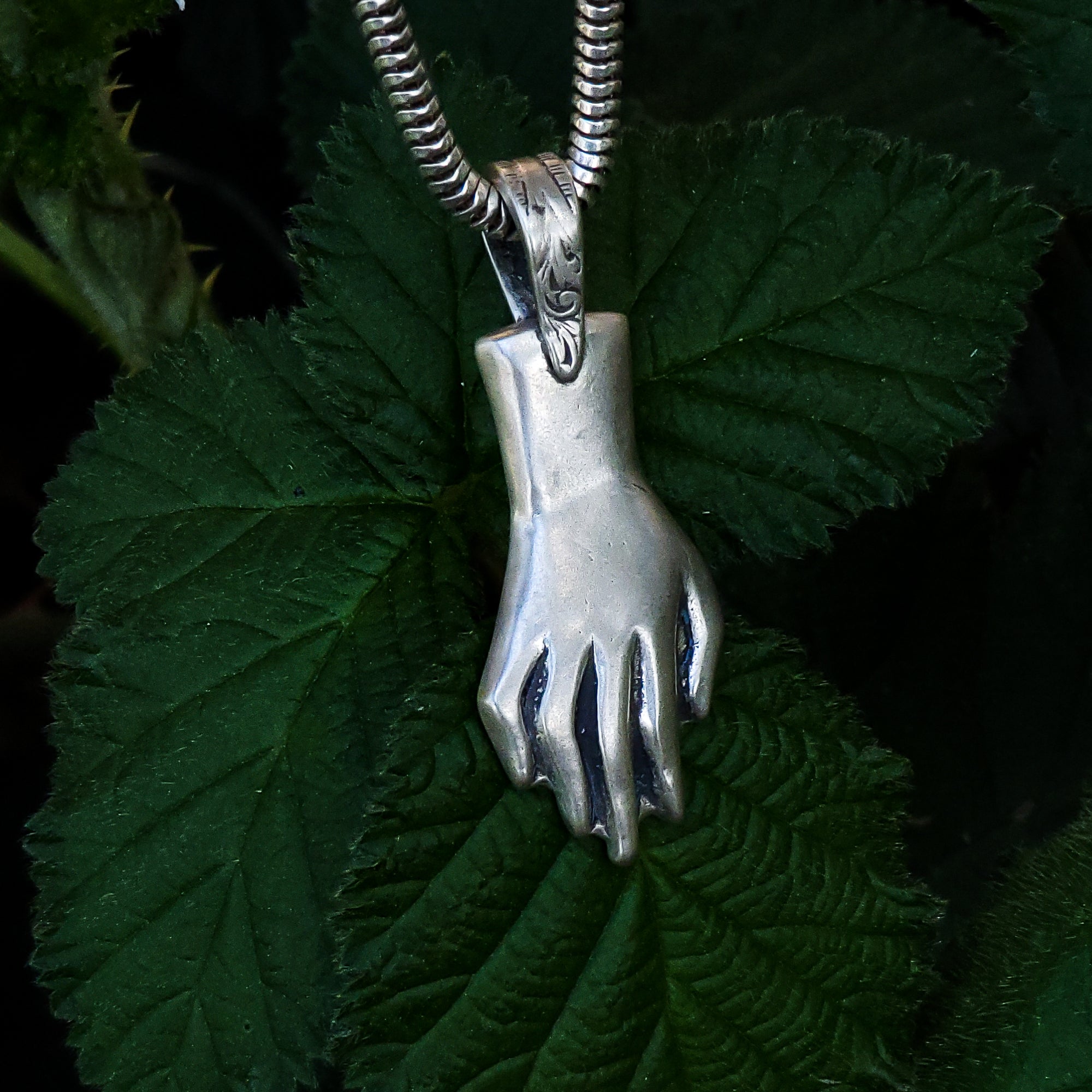 In this photo, a sterling silver pendant in the shape of a hand is placed on top of dark green leaves. The pendant is intricately detailed and beautifully crafted, with outstretched fingers that suggest a sense of openness and grace. The leaves underneath the pendant provide a contrasting backdrop that emphasizes the pendant's shimmering surface.