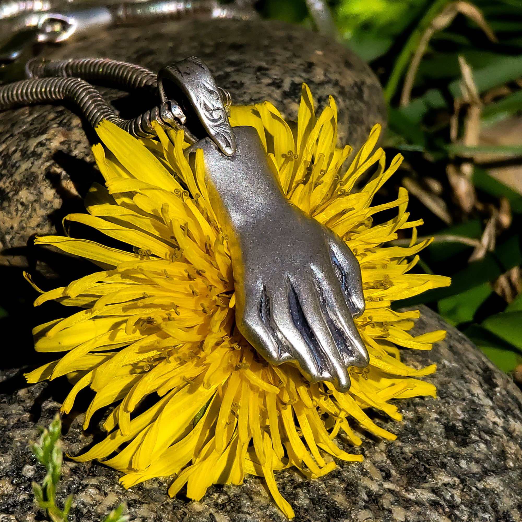 In this photo, a sterling silver pendant in the shape of a hand is resting on top of a dandelion. The pendant is beautifully crafted with intricate details. Behind the pendant, there are various sized rocks and tufts of green grass that complement the natural feel of the image. The dandelion is in full bloom and has a soft, ethereal quality that is accentuated by the shallow depth of field.