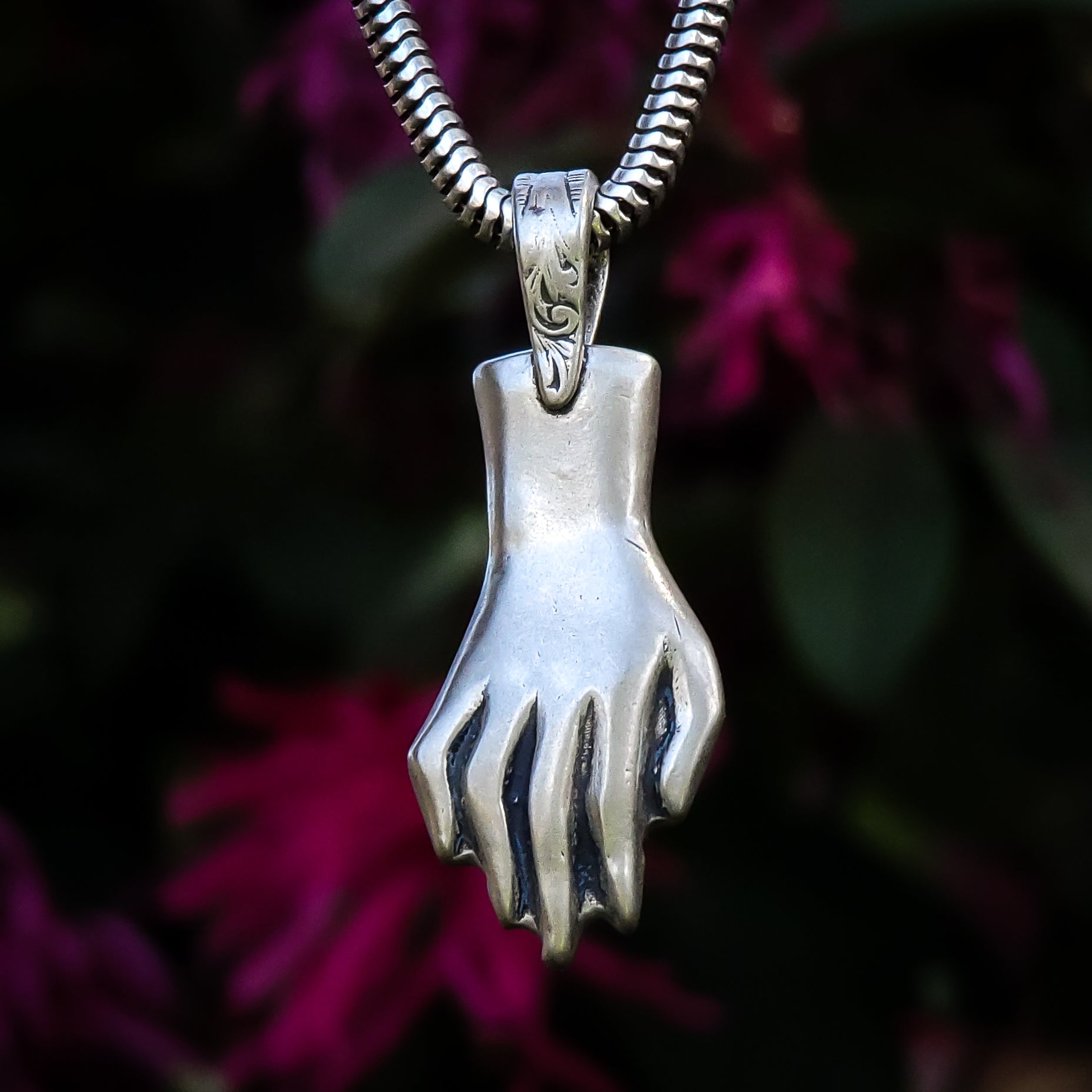 In this photo, a sterling silver pendant in the shape of a hand is suspended in front of a flowering tree. The pendant is delicately detailed, with outstretched fingers that suggest a sense of openness and welcome. It is positioned in the center of the frame, with the tree in the background, slightly out of focus. The pendant stands out against the blurred background, gleaming in the light and casting delicate shadows on the surface beneath it.
