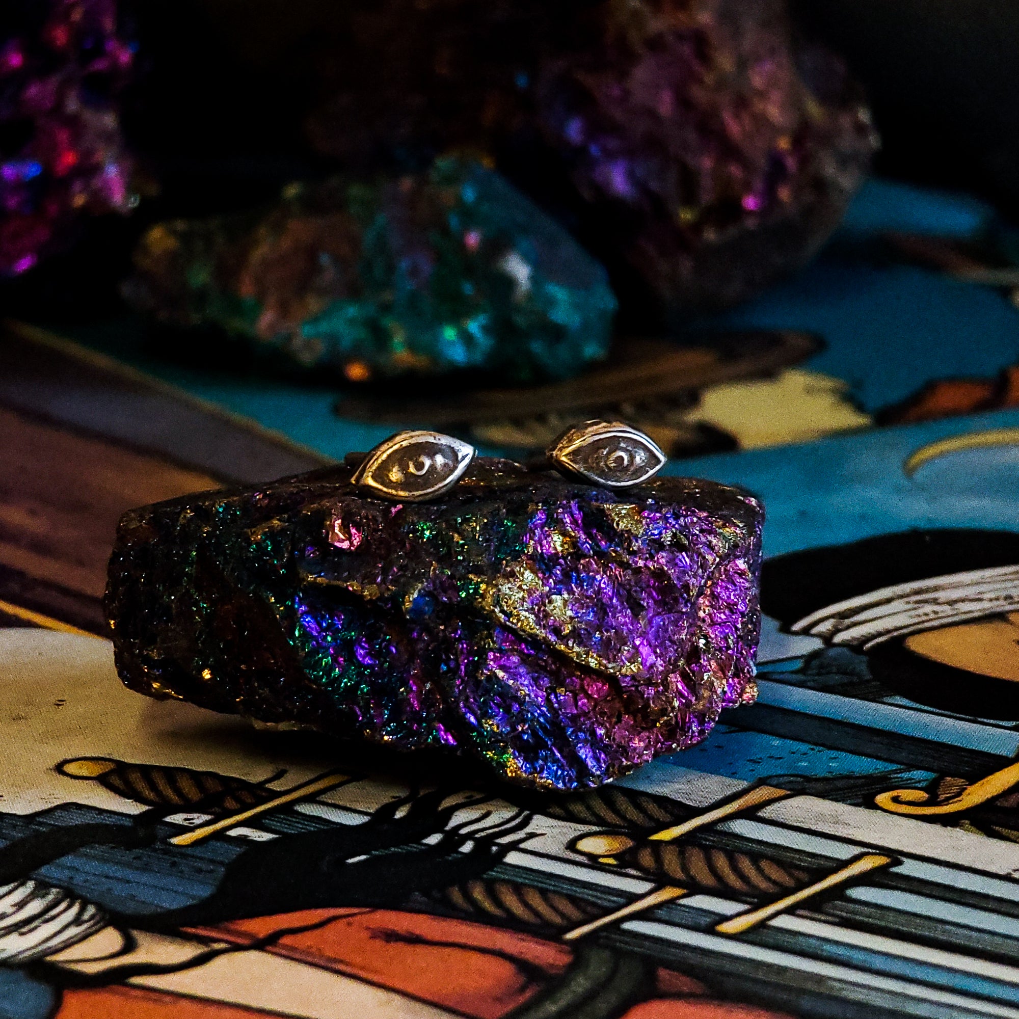 A pair of sterling silver stud earrings in the shape of human eyes sit stop a sparkly multicolored rock. Beneath the rock, there are tarot cards showing images of people with cloths over their eyes. In the background there are more multicolored rocks.